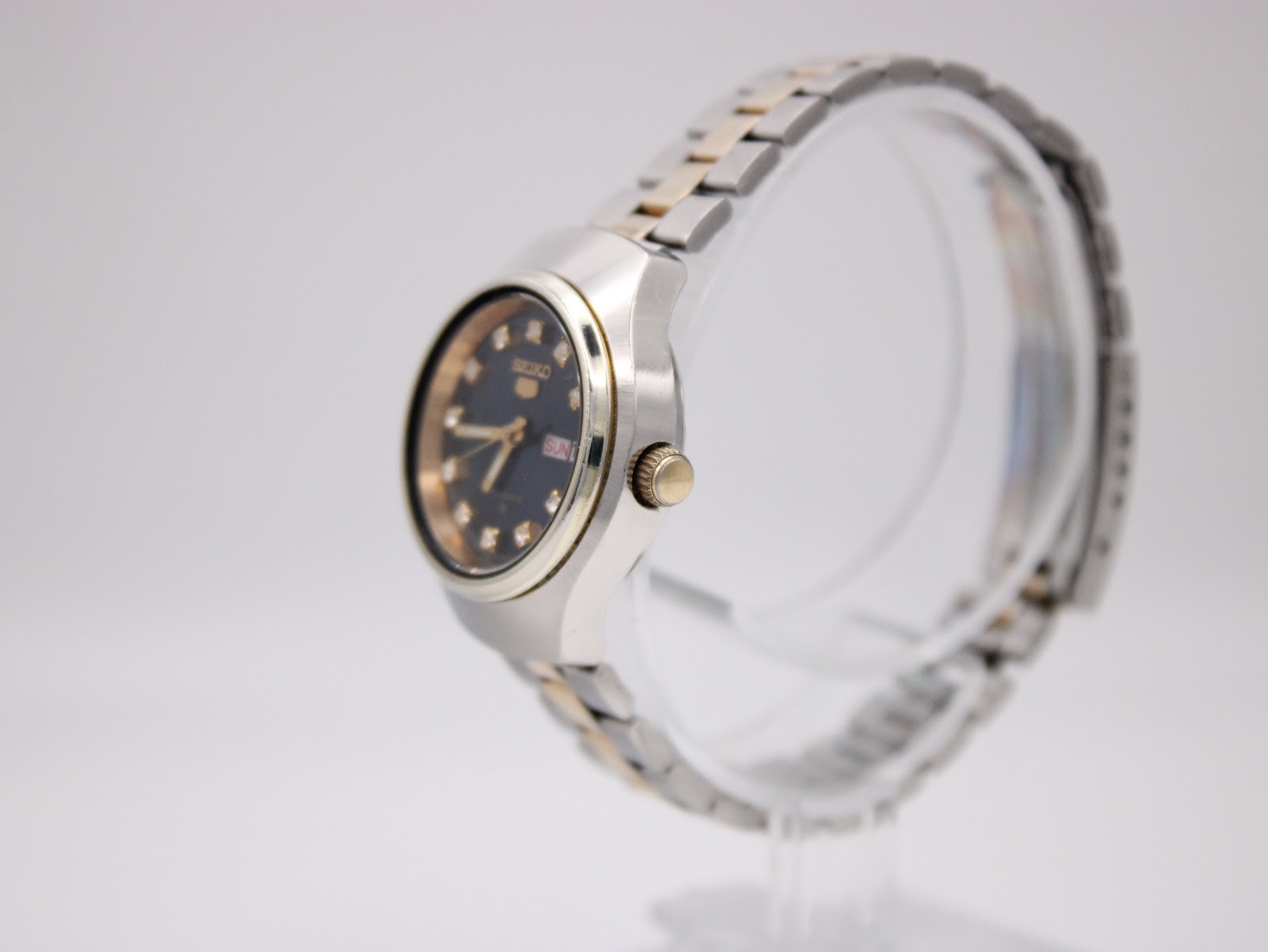 Side profile view of the Seiko watch, showing the gold-tone crown and fluted bezel detail on a clear stand.