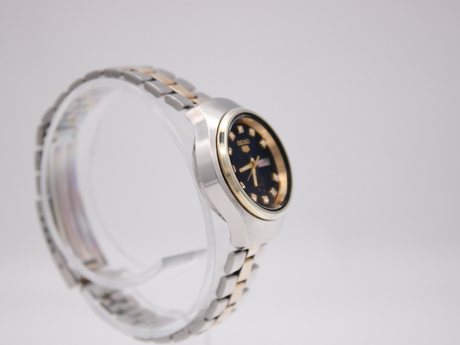 Opposite side profile view of the Seiko watch, highlighting the case shape and two-tone bracelet on a clear stand.