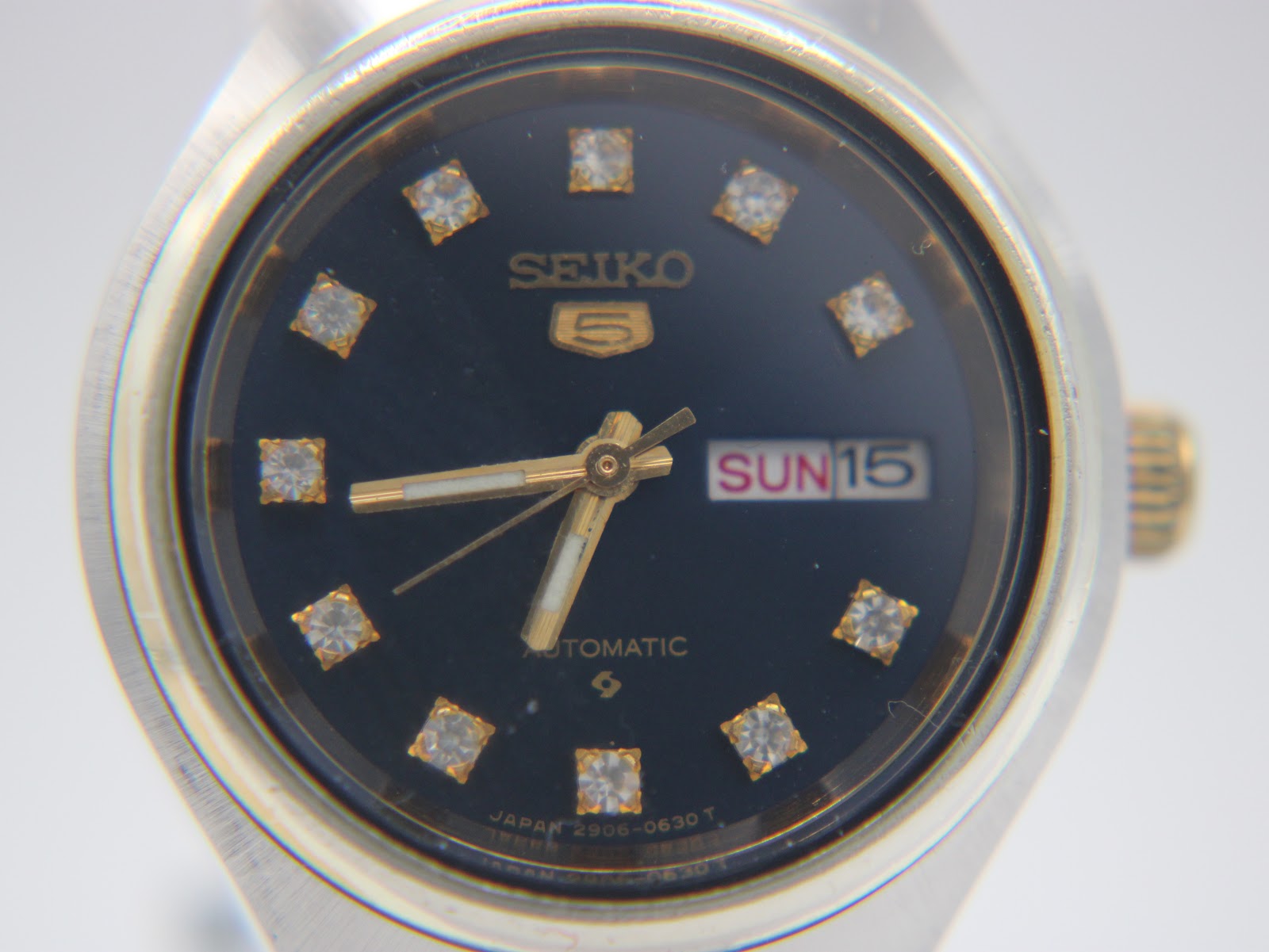 Close-up front view of the Seiko 5 black dial with gold-tone hands, stone markers, and day-date window.
