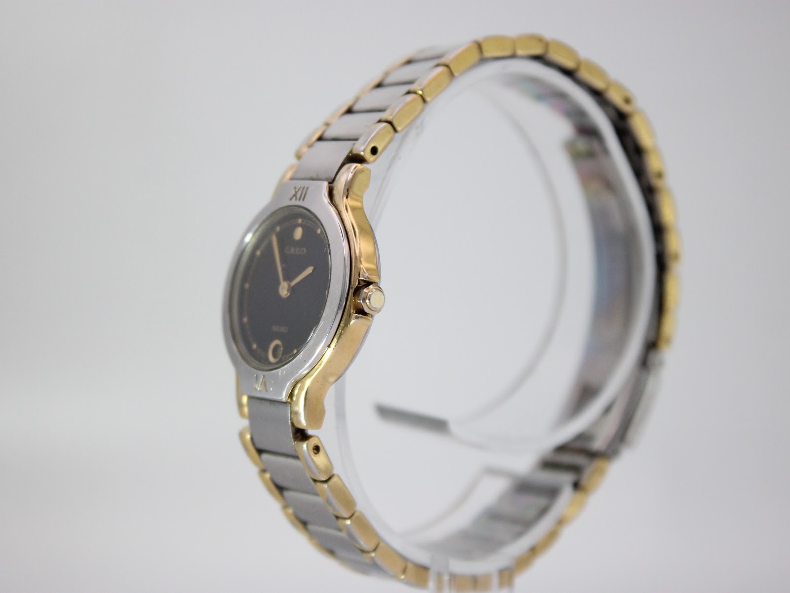 Angled side view of the Seiko Creo watch showing the gold-tone case and crown detail on a clear display stand.