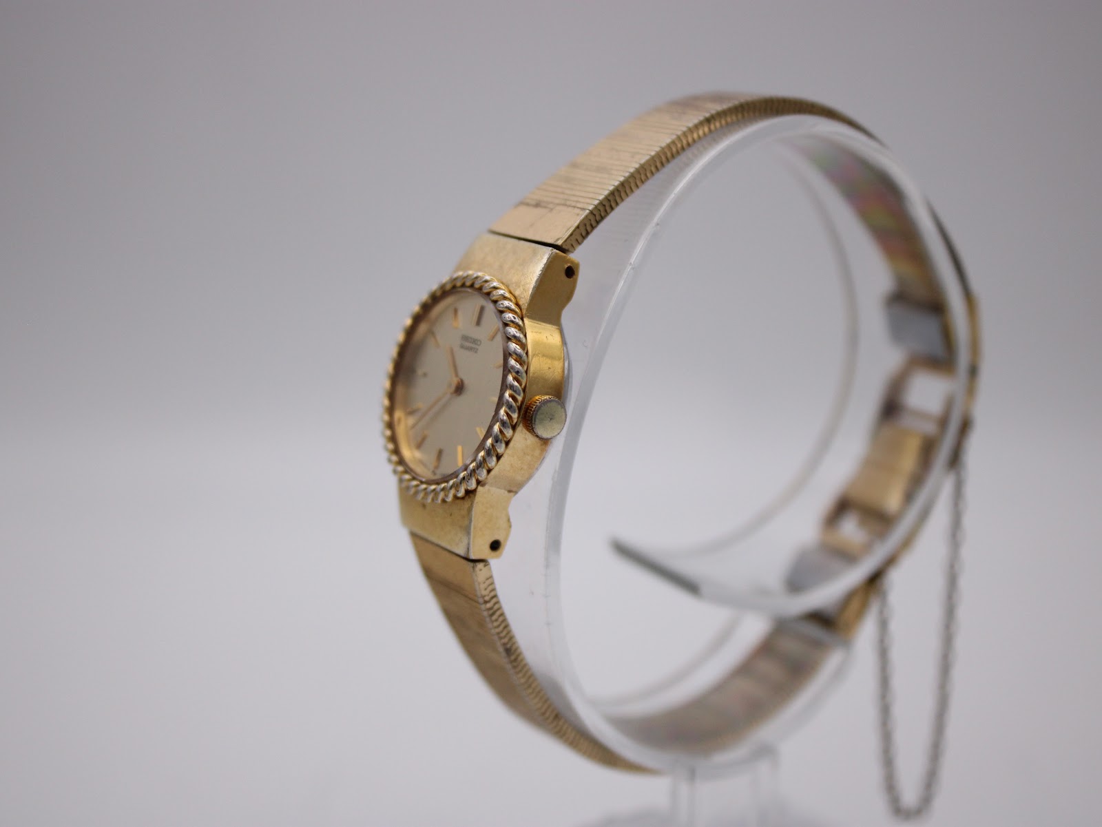 Side profile view of the gold-tone Seiko watch on a clear stand, showing the case and textured mesh bracelet.