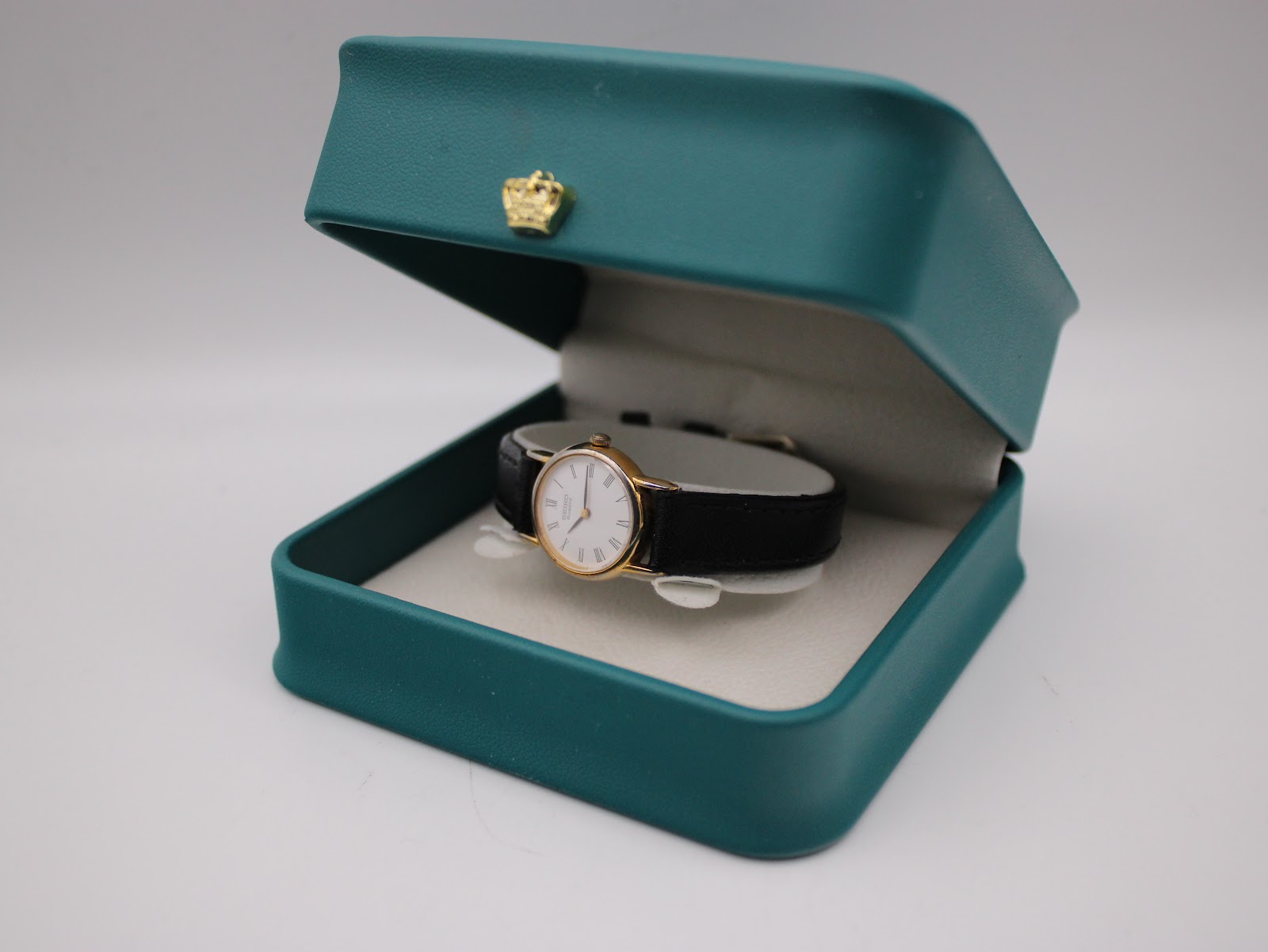 The Seiko watch with its black leather strap displayed within an open teal coloured presentation box with a gold crown emblem.