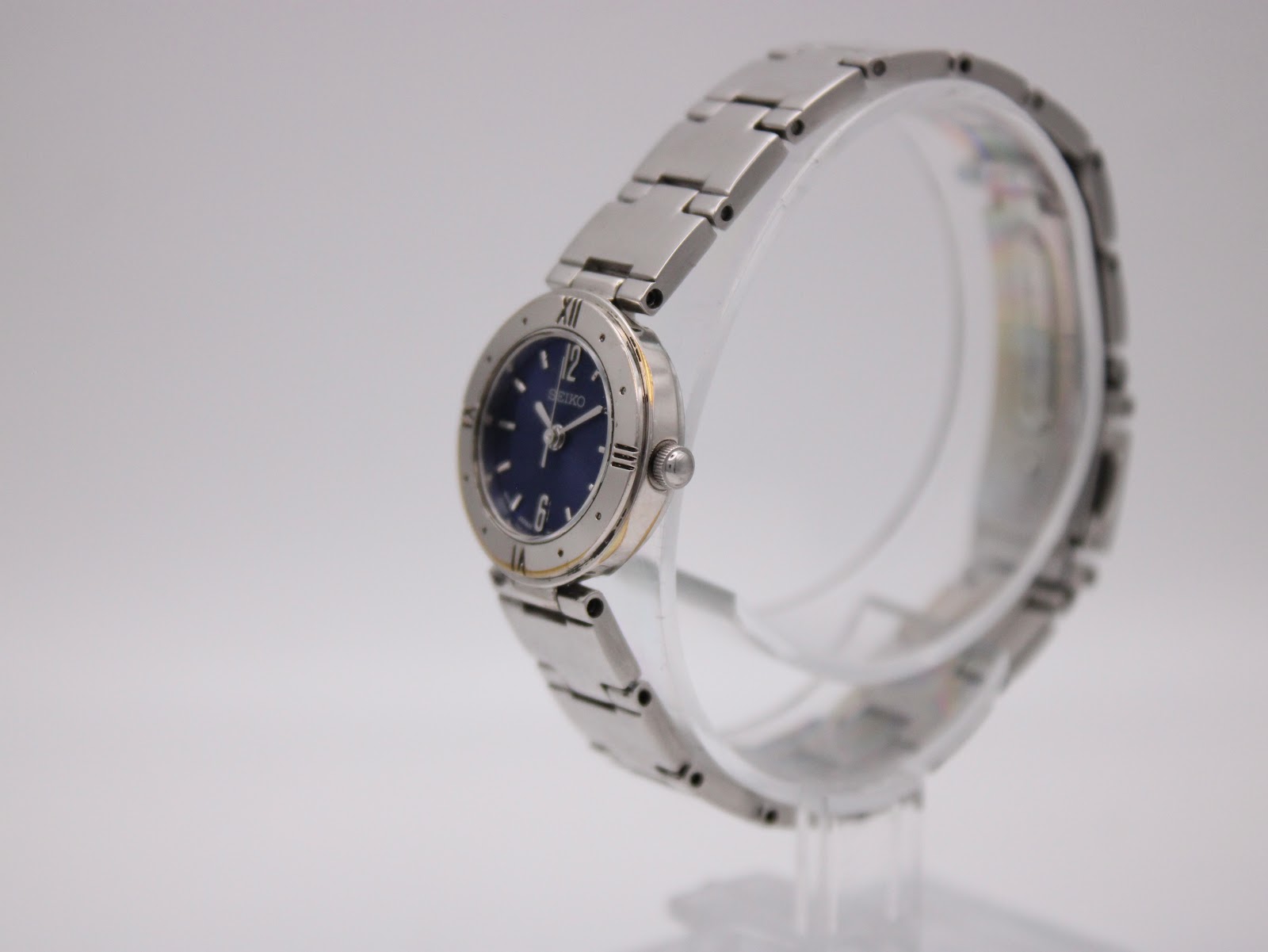 Angled side view of the Seiko watch, showing the silver-tone case and integrated bracelet on a clear display stand.