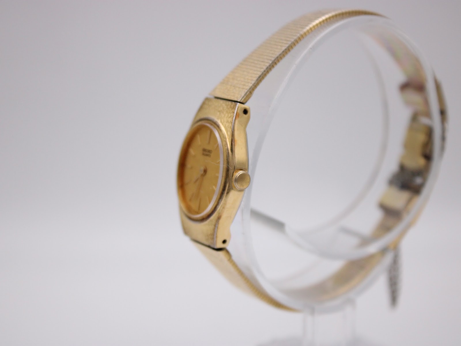 Angled side view of the gold Seiko watch showing the crown and case profile on a transparent display stand.