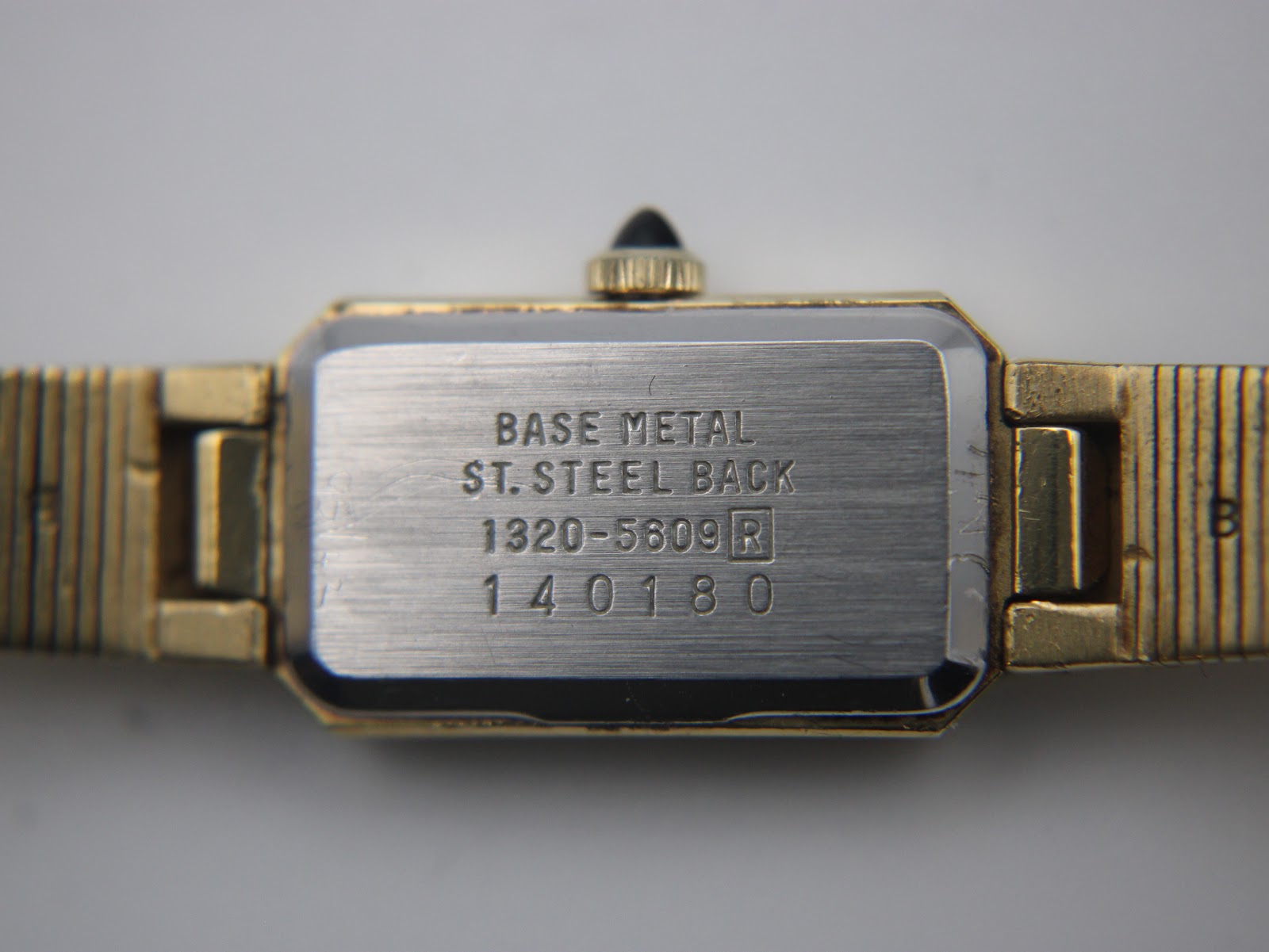 Close-up of the watch case back, revealing engravings 'BASE METAL', 'ST. STEEL BACK', model '1320-5609 R', and serial number '140180'.