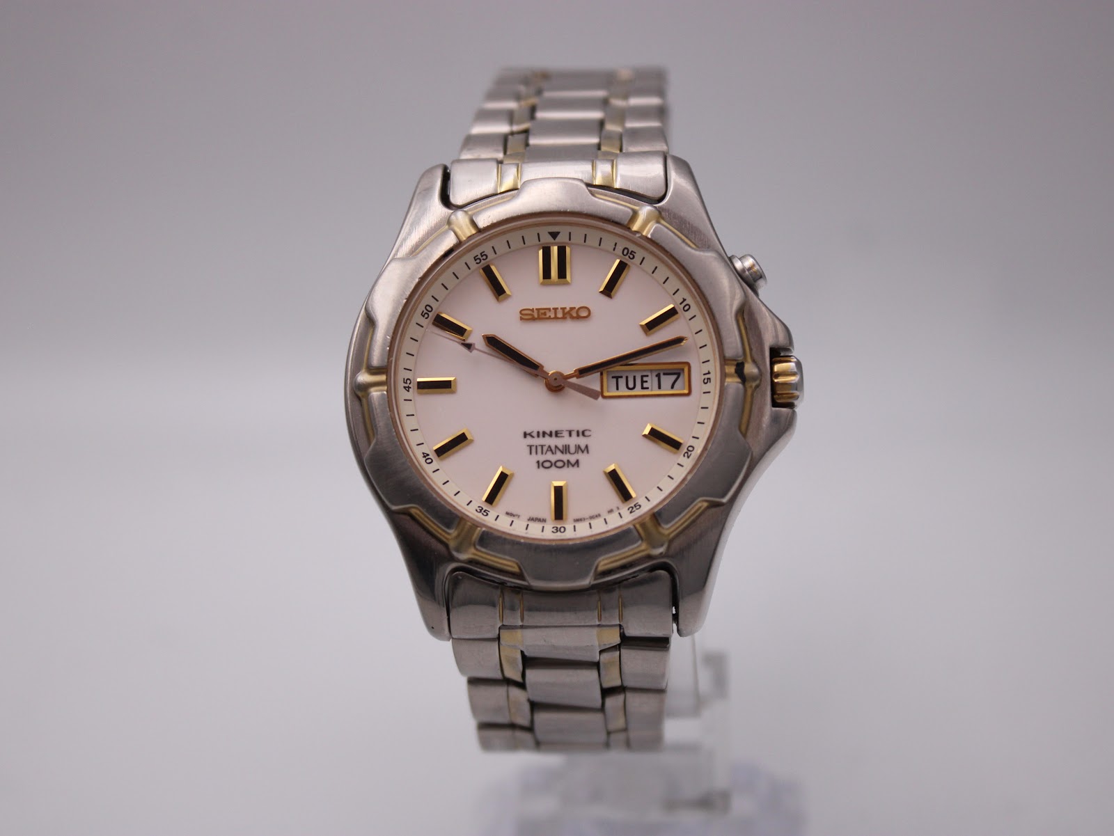 Clear front view of the two-tone Seiko Kinetic titanium watch, with its white dial, gold baton indices, and day-date window.
