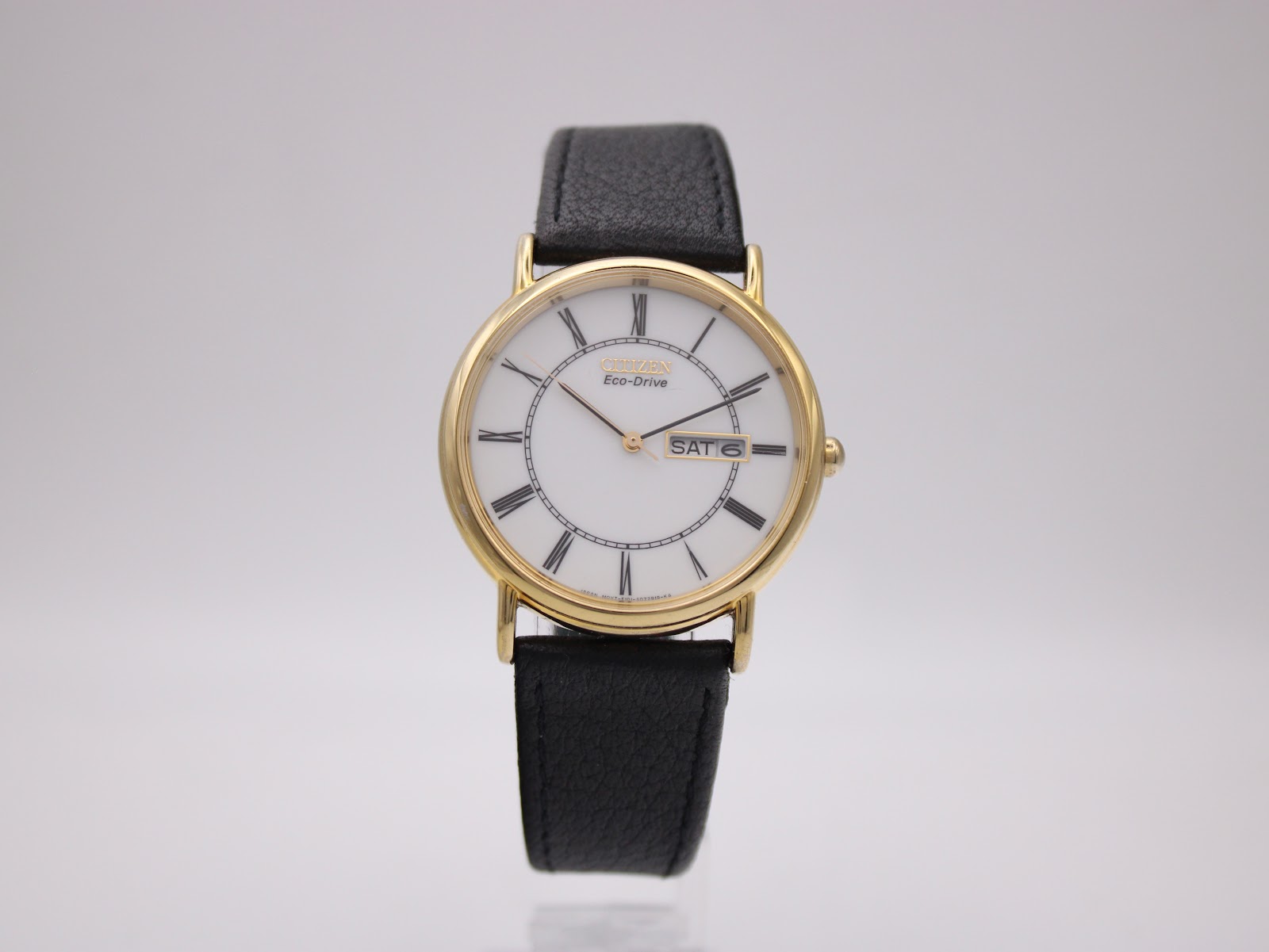 Frontal view of the Citizen Eco-Drive watch on a clear stand, showing white dial, gold case, and black leather strap.