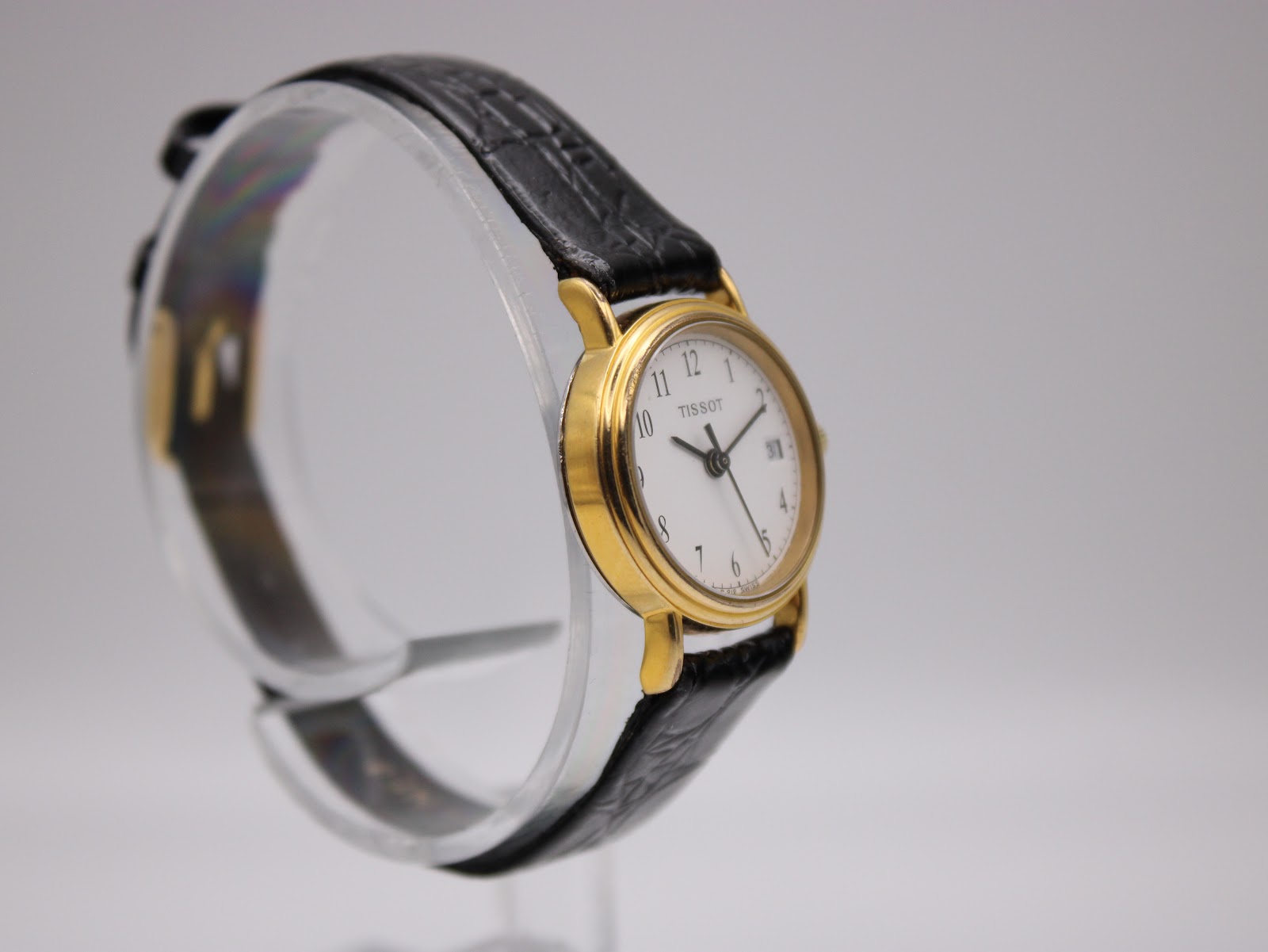 Angled left-side view of the Tissot C 212 watch on a display stand, showcasing the side profile of the gold-tone case and black leather strap.