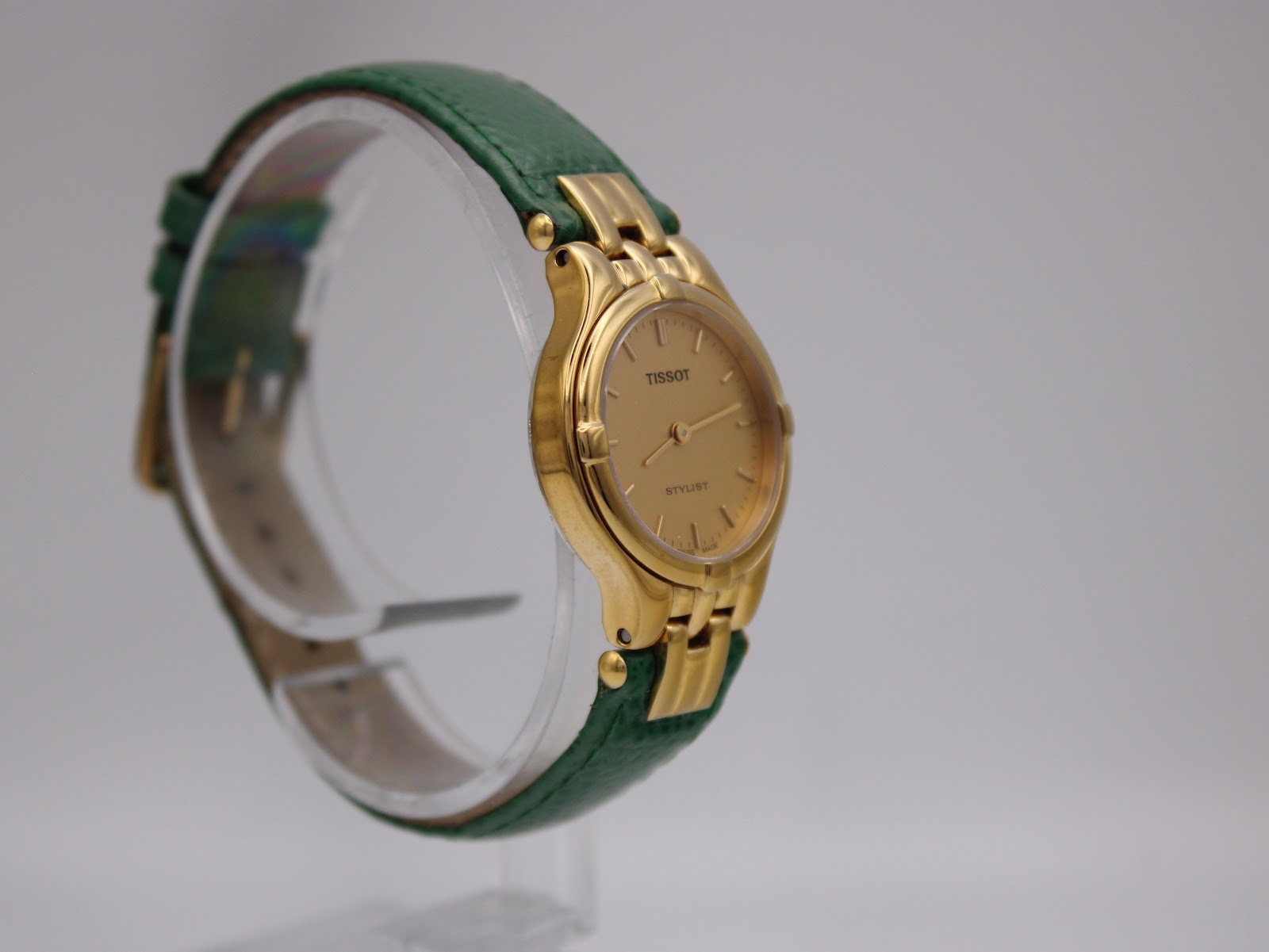 Side profile of the Tissot Stylist watch on a clear display stand, showing the strap's texture and integrated gold links.