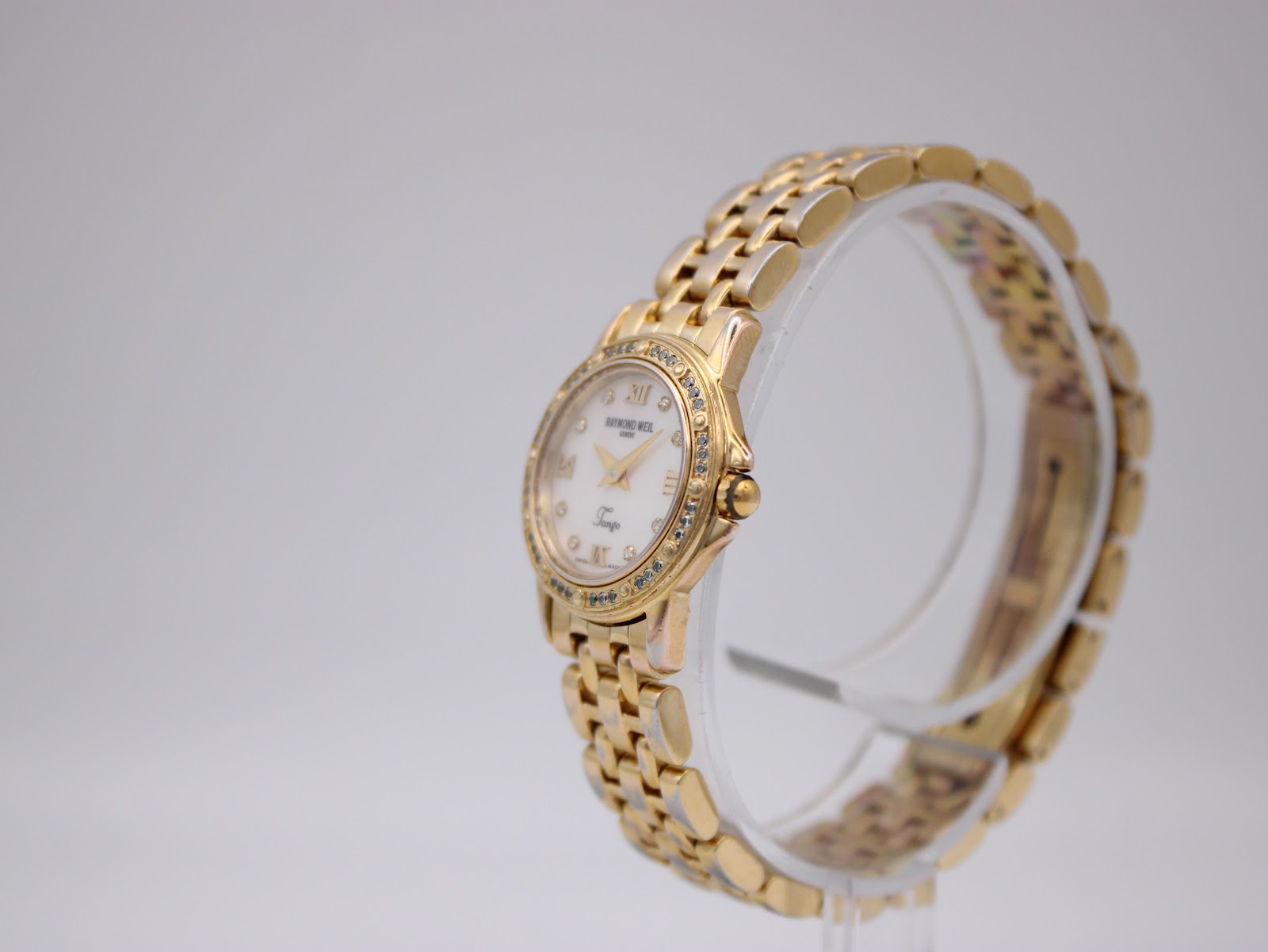 Angled side view of the Raymond Weil Tango watch on a clear stand, highlighting the gold-plated case and bracelet.
