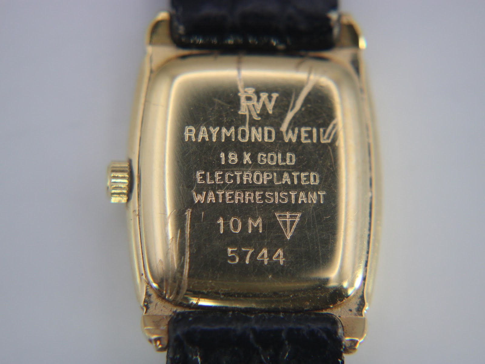 Close-up of the watch case back, revealing engravings including 'RW', 'RAYMOND WEIL', '18 K GOLD ELECTROPLATED', 'WATERRESISTANT', '10 M', and '5744'.