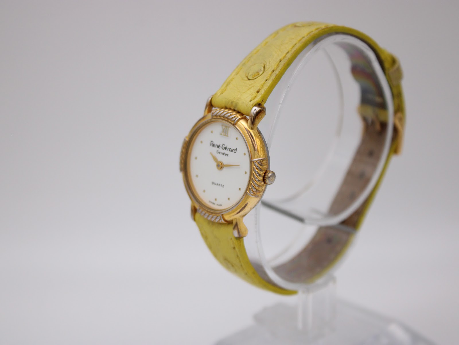 Angled view of the René Gérard watch, highlighting the ridged gold-toned bezel and a textured yellow leather strap on a clear stand.