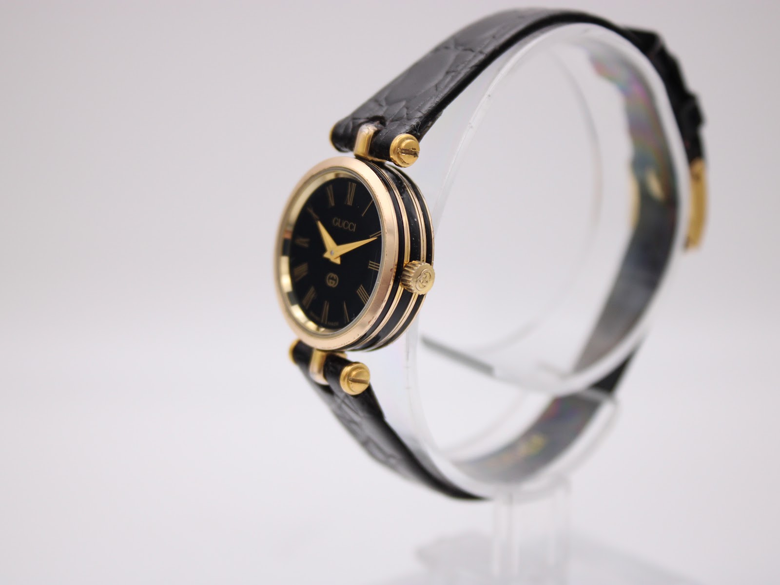 Angled side view of the Gucci 2000L watch on a clear display stand, highlighting the ribbed gold-tone case and crown.