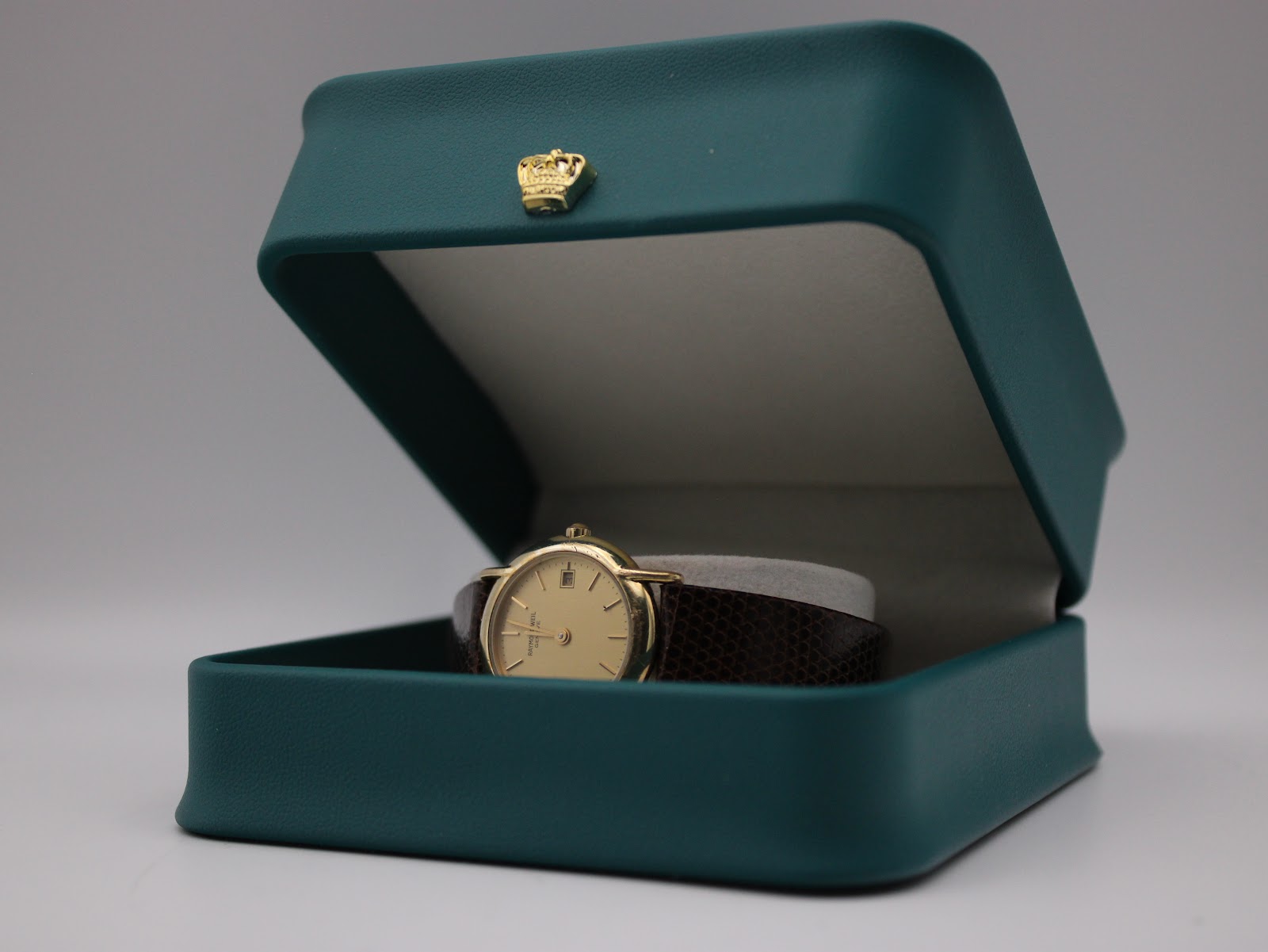 The Raymond Weil watch placed inside a generic teal display box with a gold crown emblem, suggesting presentation.