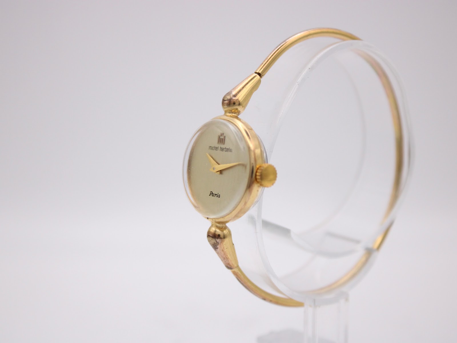 Three-quarter view of the Michel Herbelin watch showing the gold-tone case, cream dial, and integrated bangle bracelet on a clear stand.