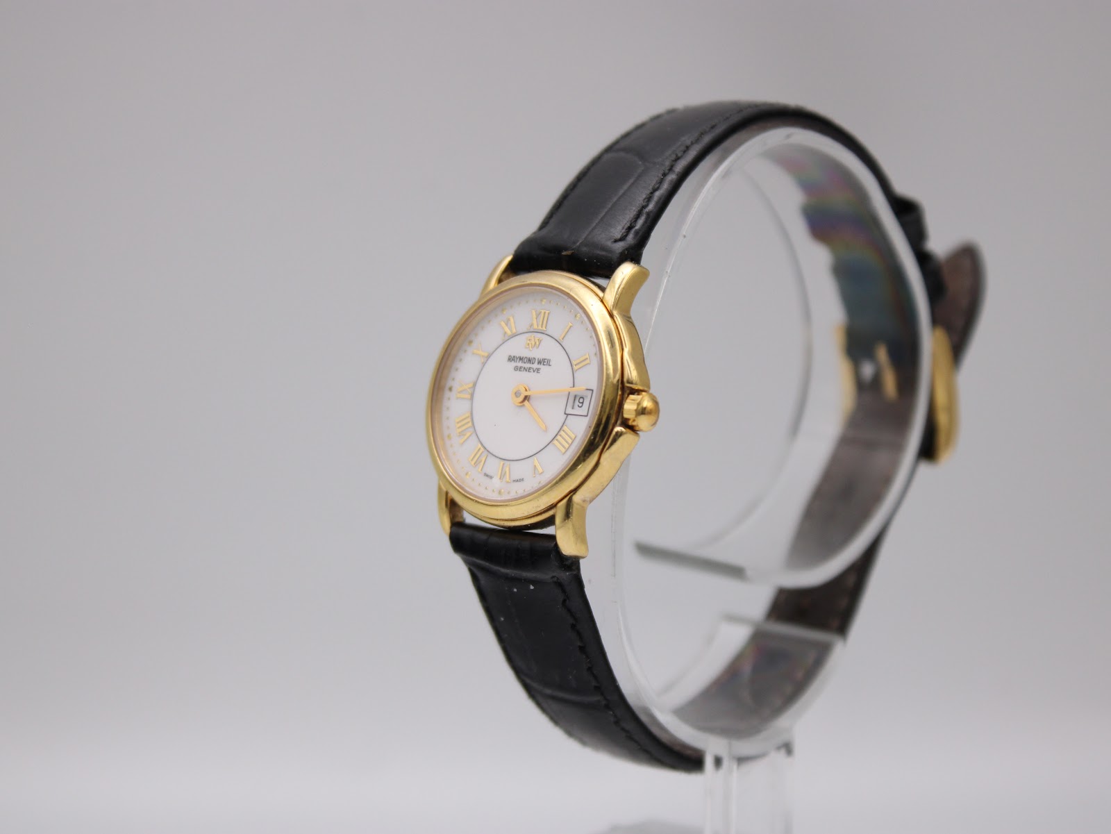 Angled view of the Raymond Weil 9972 watch on a clear stand, showcasing the gold plated case and crown, with the black leather strap curving.