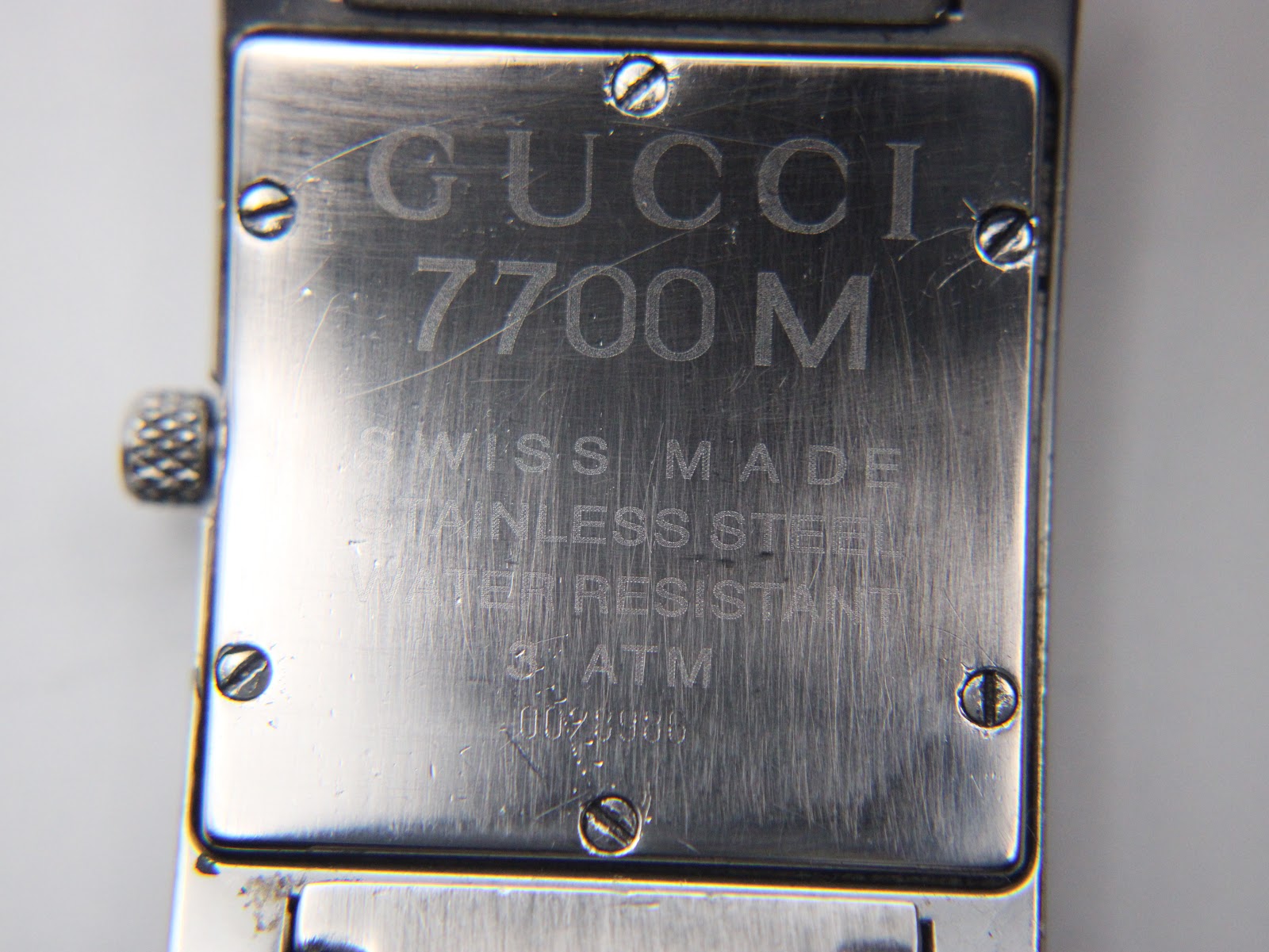 Close-up of the watch case back, clearly showing "GUCCI 7700 M", "SWISS MADE", "STAINLESS STEEL", "WATER RESISTANT", "3 ATM", and serial number "0028986" engravings.
