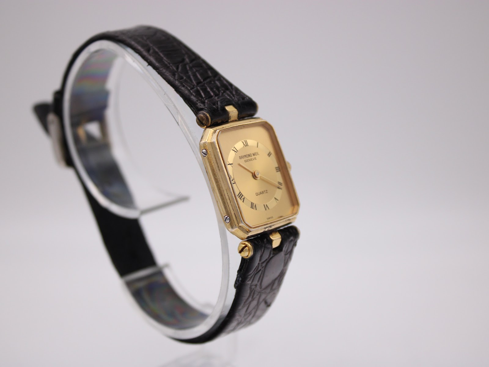 Another angled side view of the Raymond Weil watch highlighting the case shape and black leather strap on a clear stand.