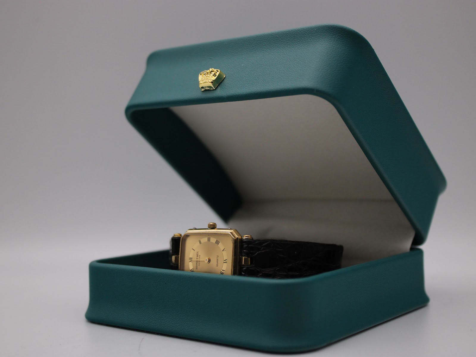 The Raymond Weil watch resting inside an open, empty teal-coloured watch display box.