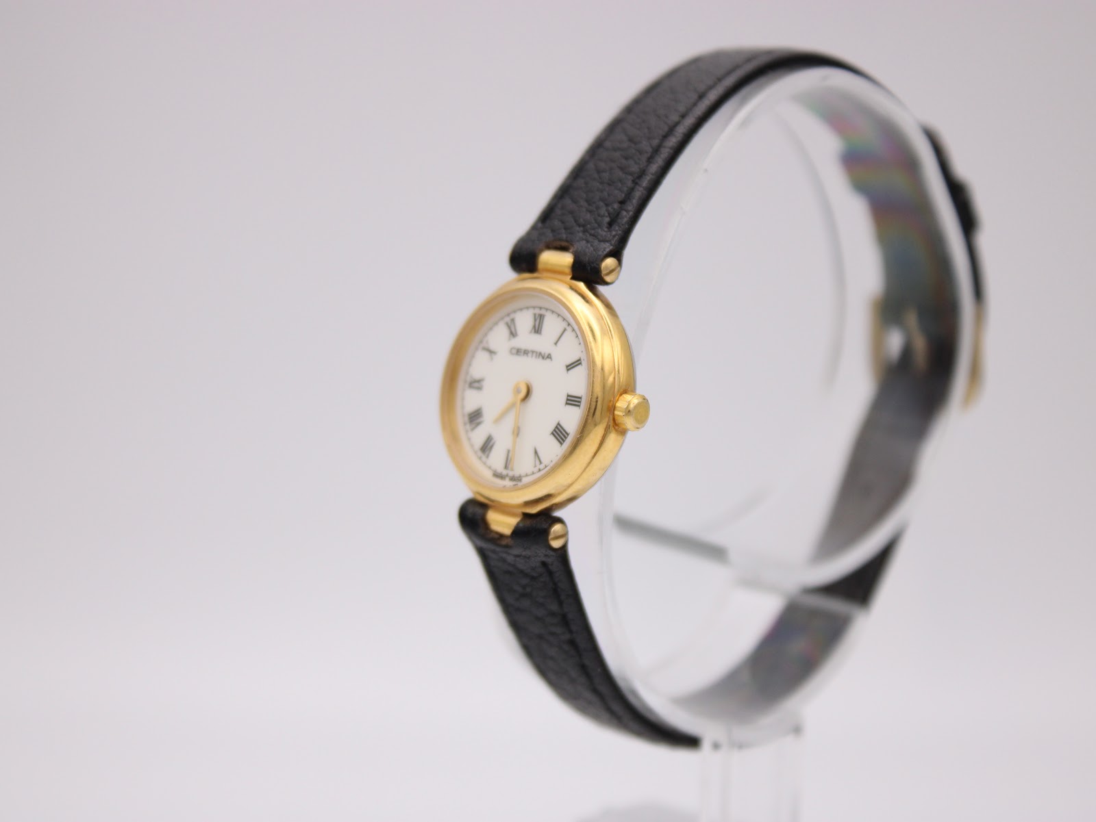 Angled left view of Certina ladies' watch on stand, showing gold case and black leather strap.