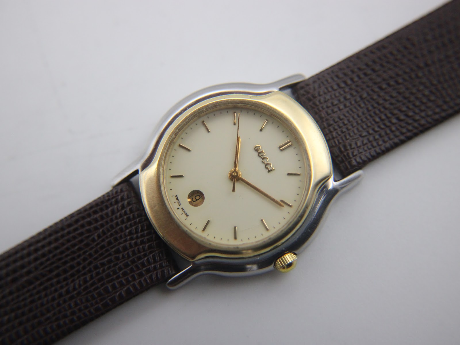 High-angle view of the Gucci watch dial, showcasing the cream face, gold hands, minimalist indices, and date window at 6 o'clock.