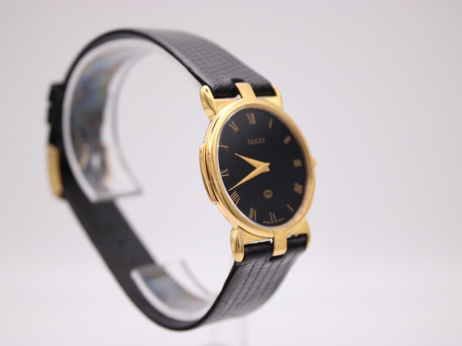 Opposite angled side view of the Gucci 3400M watch on a clear display stand, highlighting the gold case and black strap.