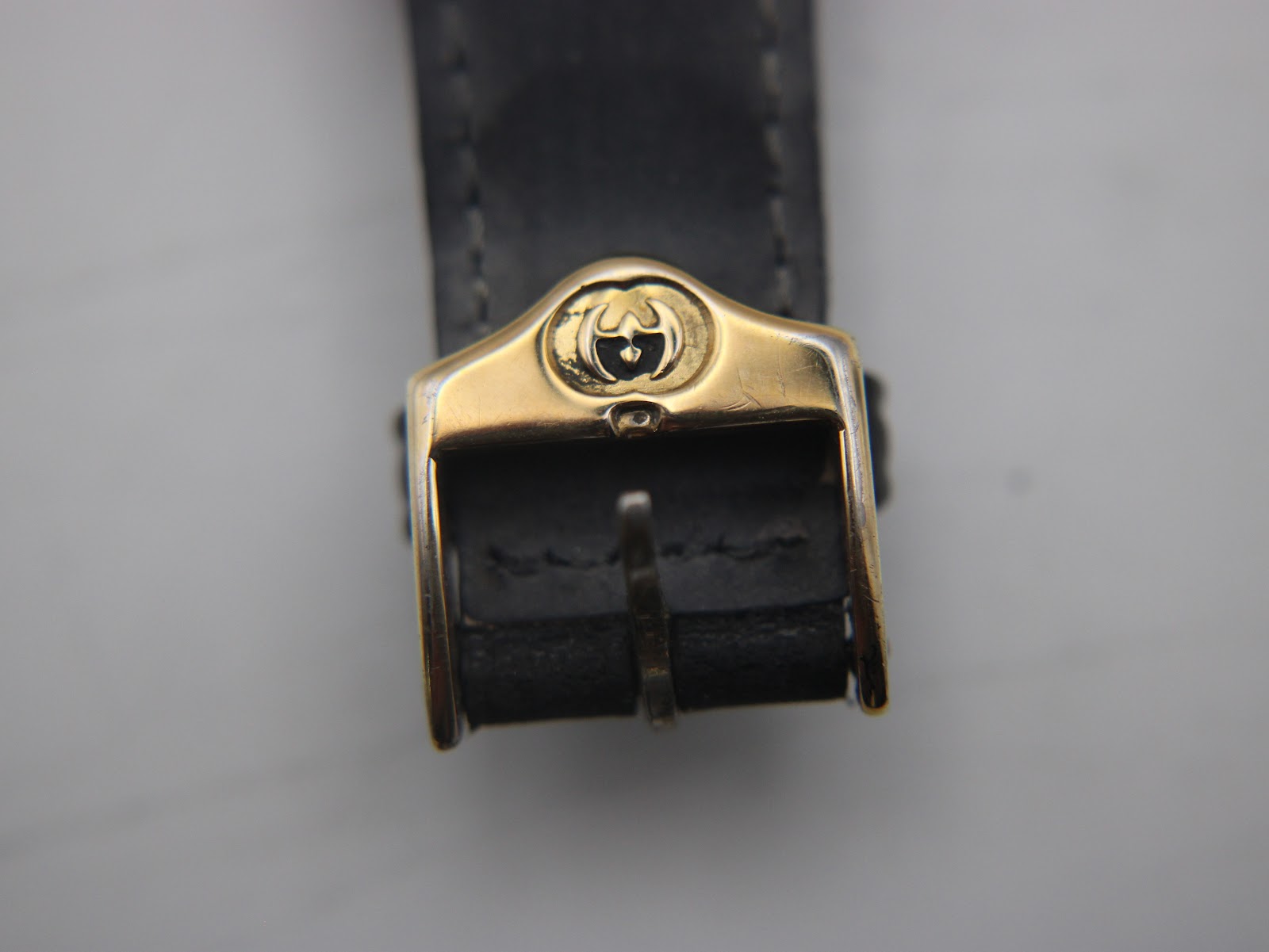 Close-up of the gold-tone buckle of the black leather strap, featuring the embossed Gucci 'G' logo.