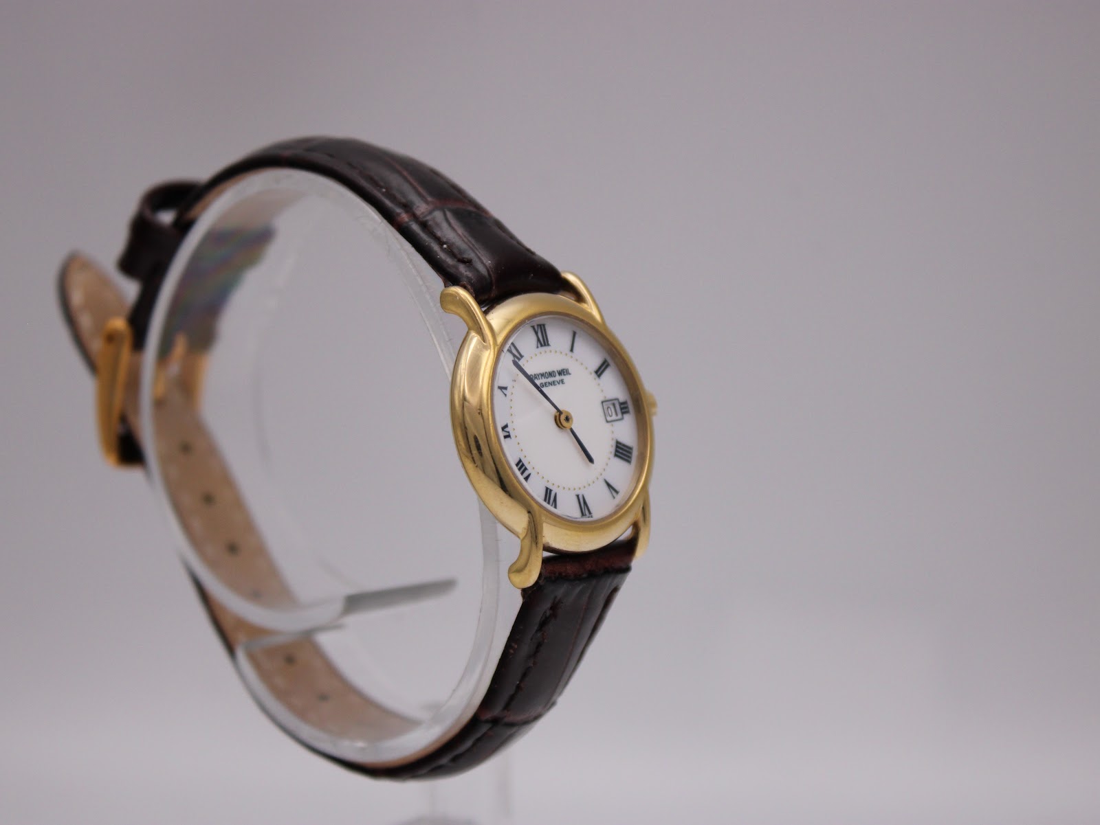 Another angled side view of the Raymond Weil watch, showing the dial, crown, and brown leather strap on a clear stand.