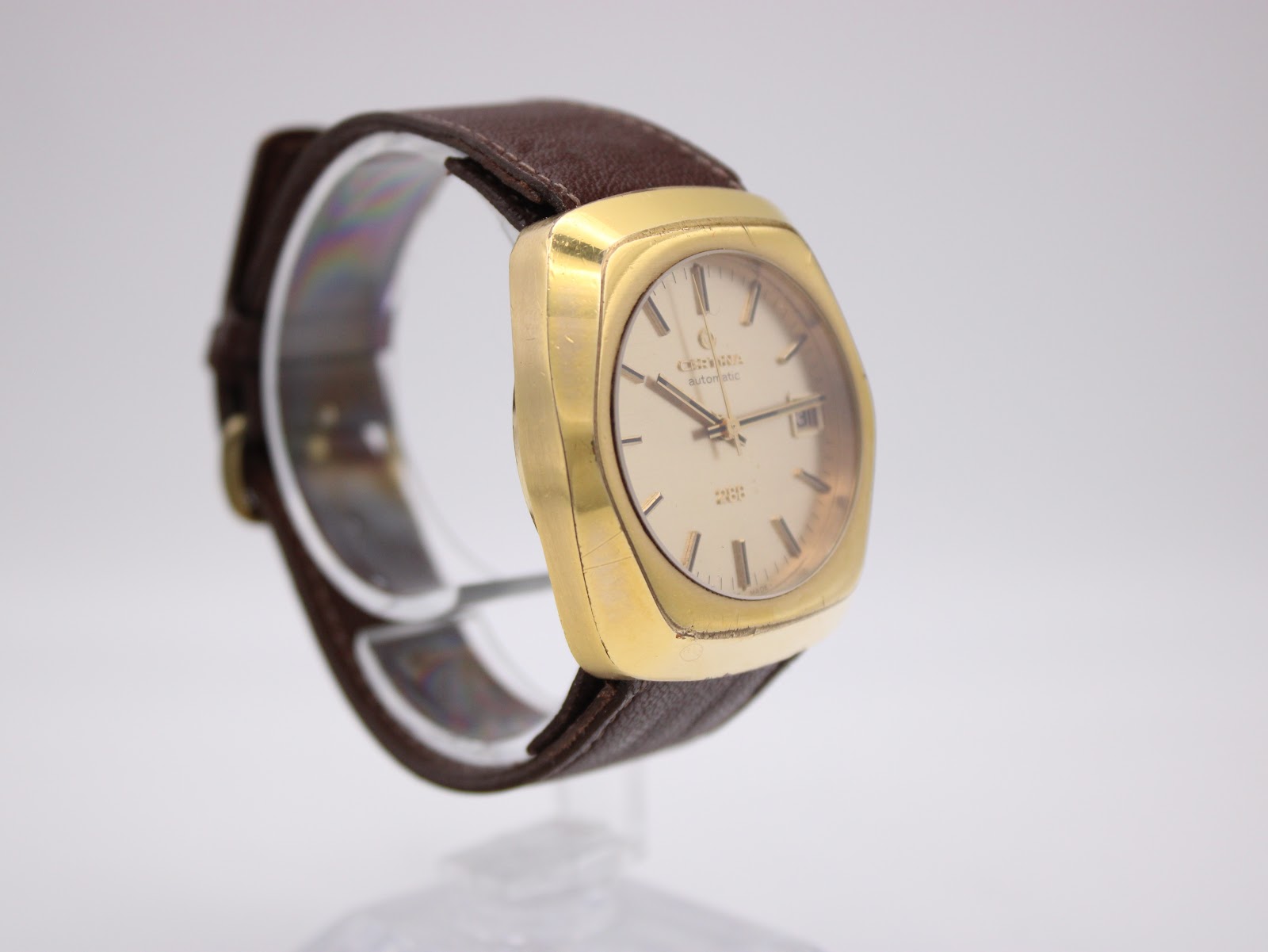 Angled side view of the Certina 288 watch, showing the left side of the gold-plated case, with the brown leather strap on a stand.