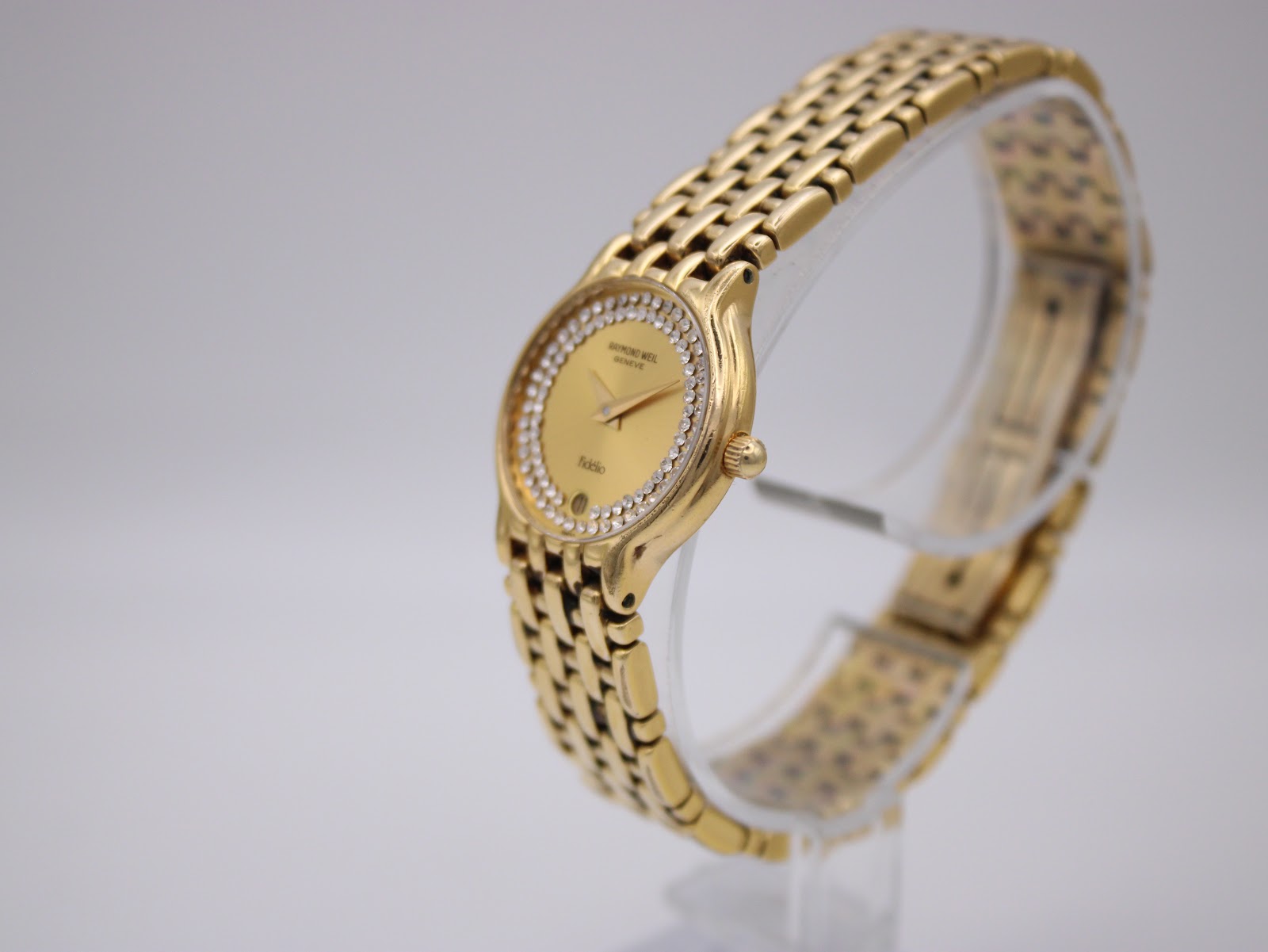 Angled side view of the Raymond Weil Fidelio gold watch showing its integrated bracelet on a clear display stand.