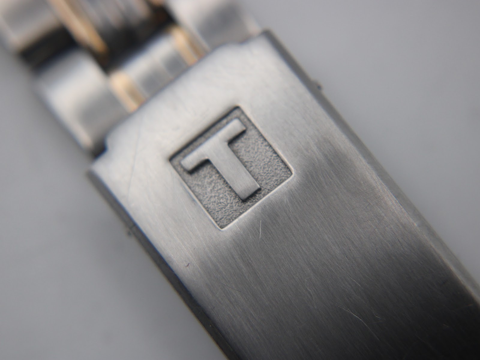 Close-up of the brushed stainless steel folding clasp of the Tissot bracelet, with a clear Tissot 'T' logo.