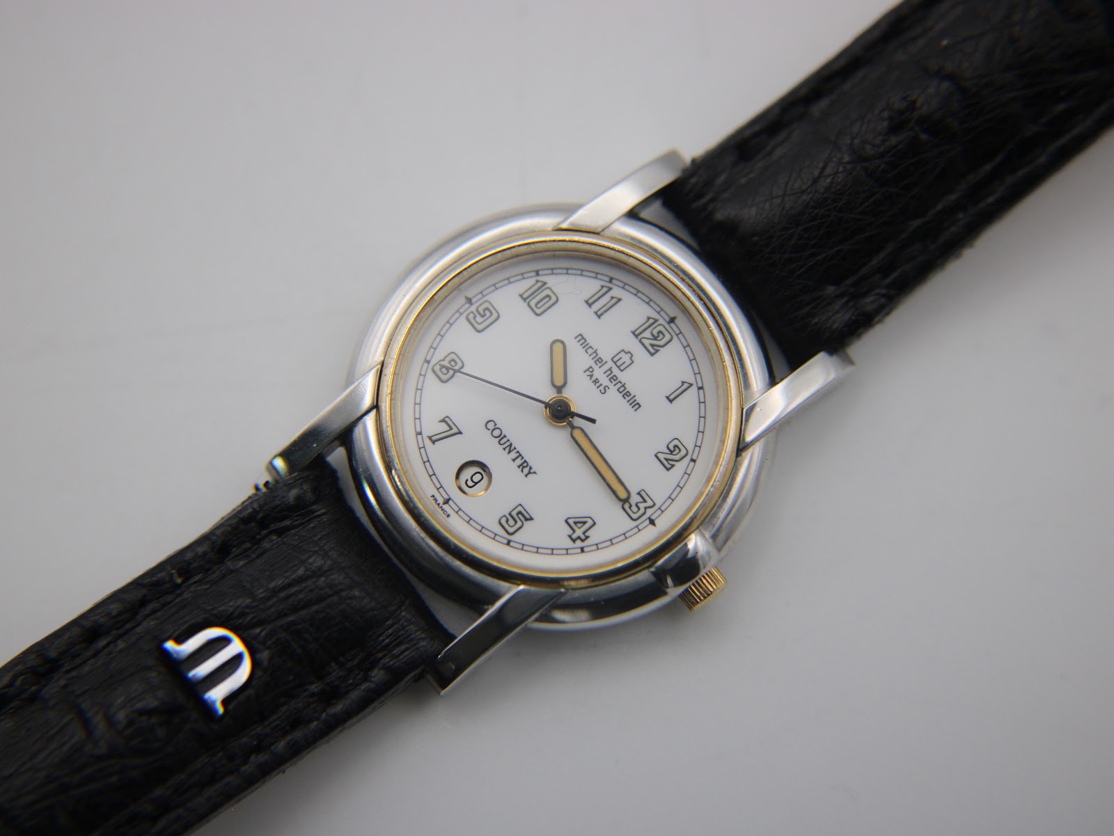 Angled front view of the Michel Herbelin watch, showcasing the white dial, gold bezel, and black leather strap with 'M' logo.
