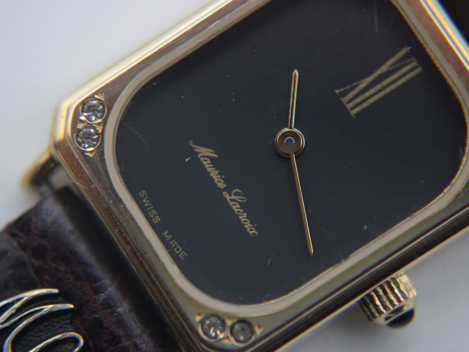 Macro close-up of the watch dial, showing the 'Maurice Lacroix' signature, Roman numeral XII, and clear crystals set into the gold-tone bezel.