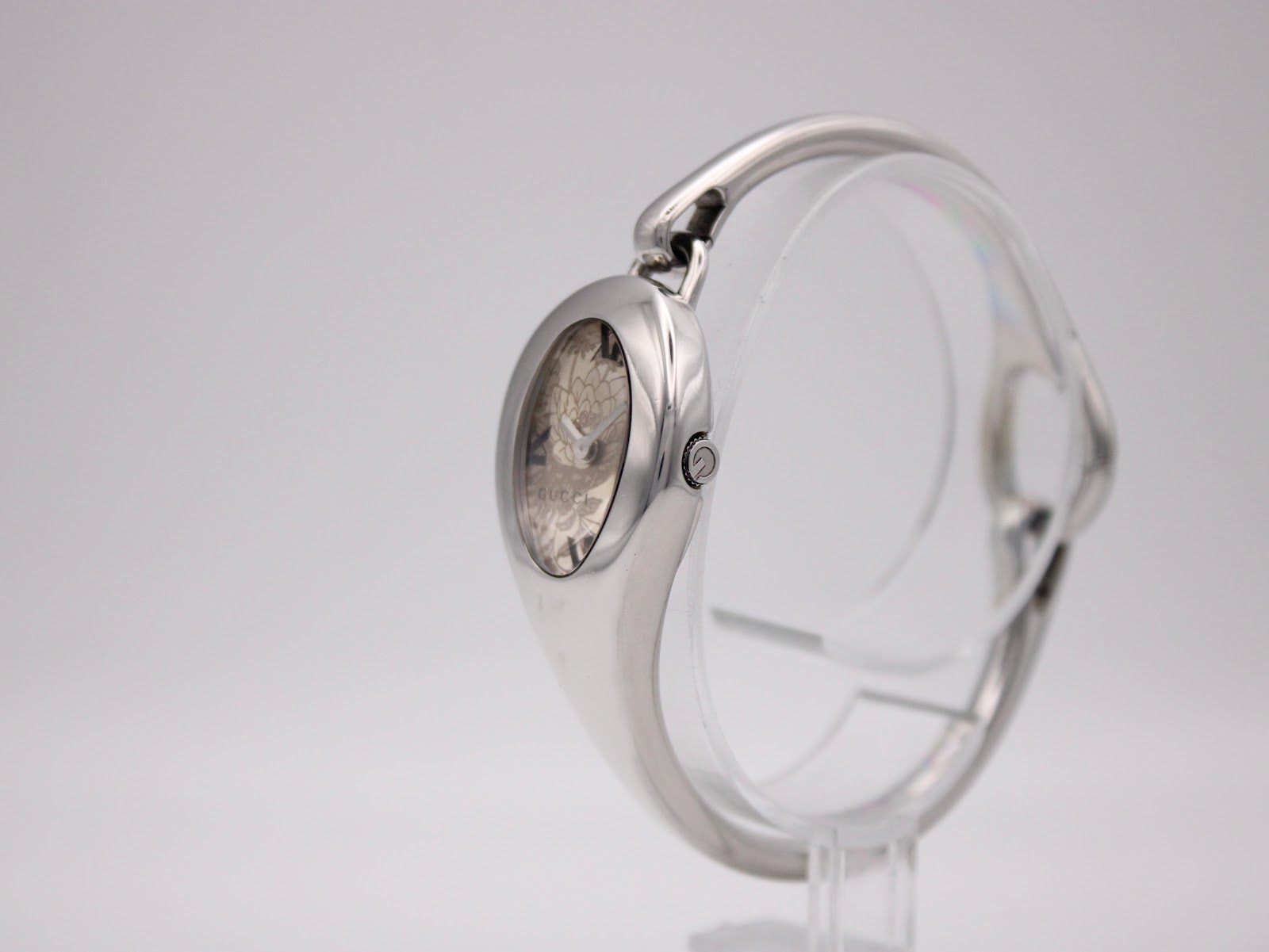 Side view of the Gucci 103 watch, showing the crown and the integrated silver bracelet, on a transparent stand.