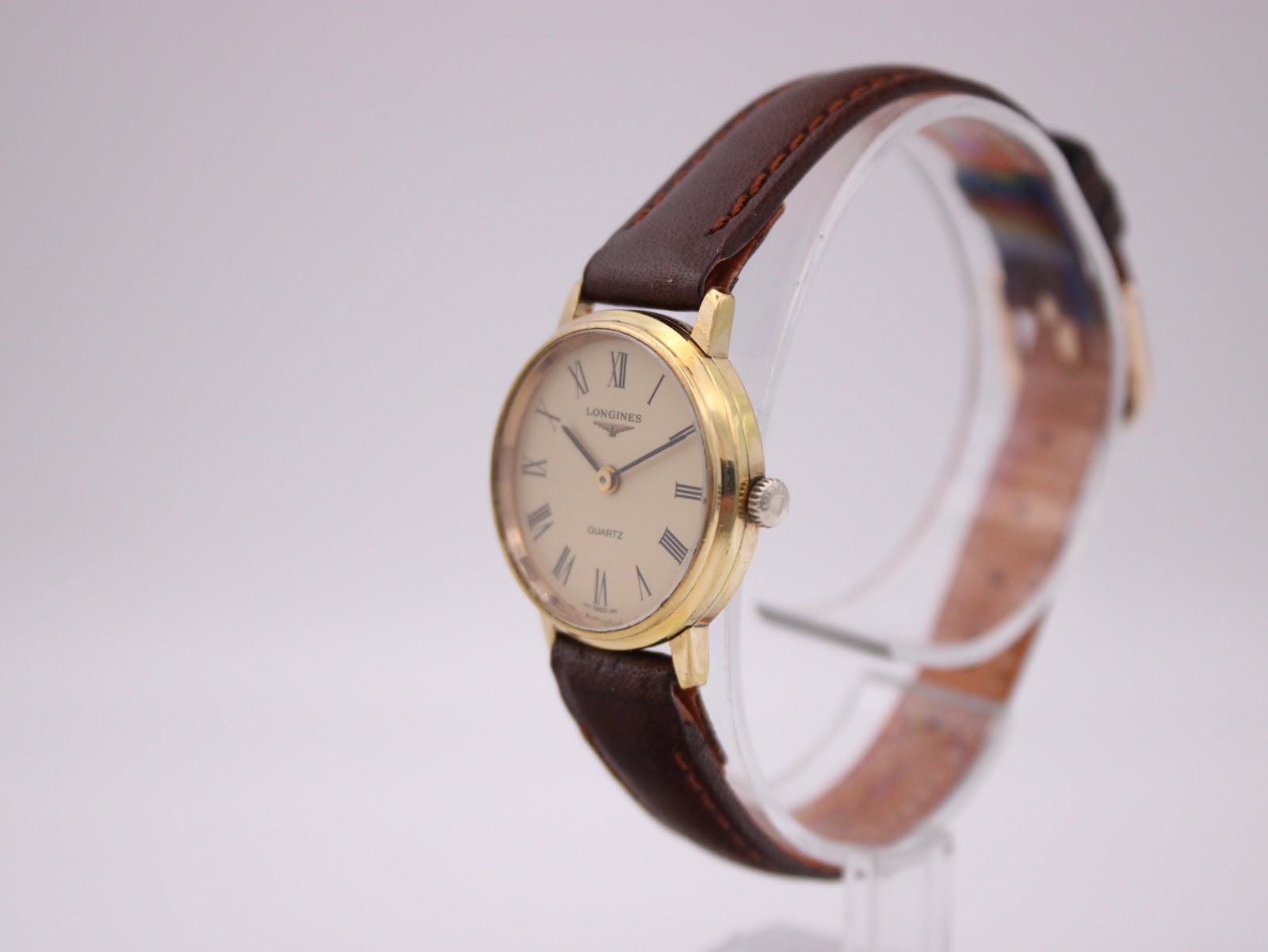Angled front view of the Longines watch on a clear display stand, showing the gold tone case and brown strap.