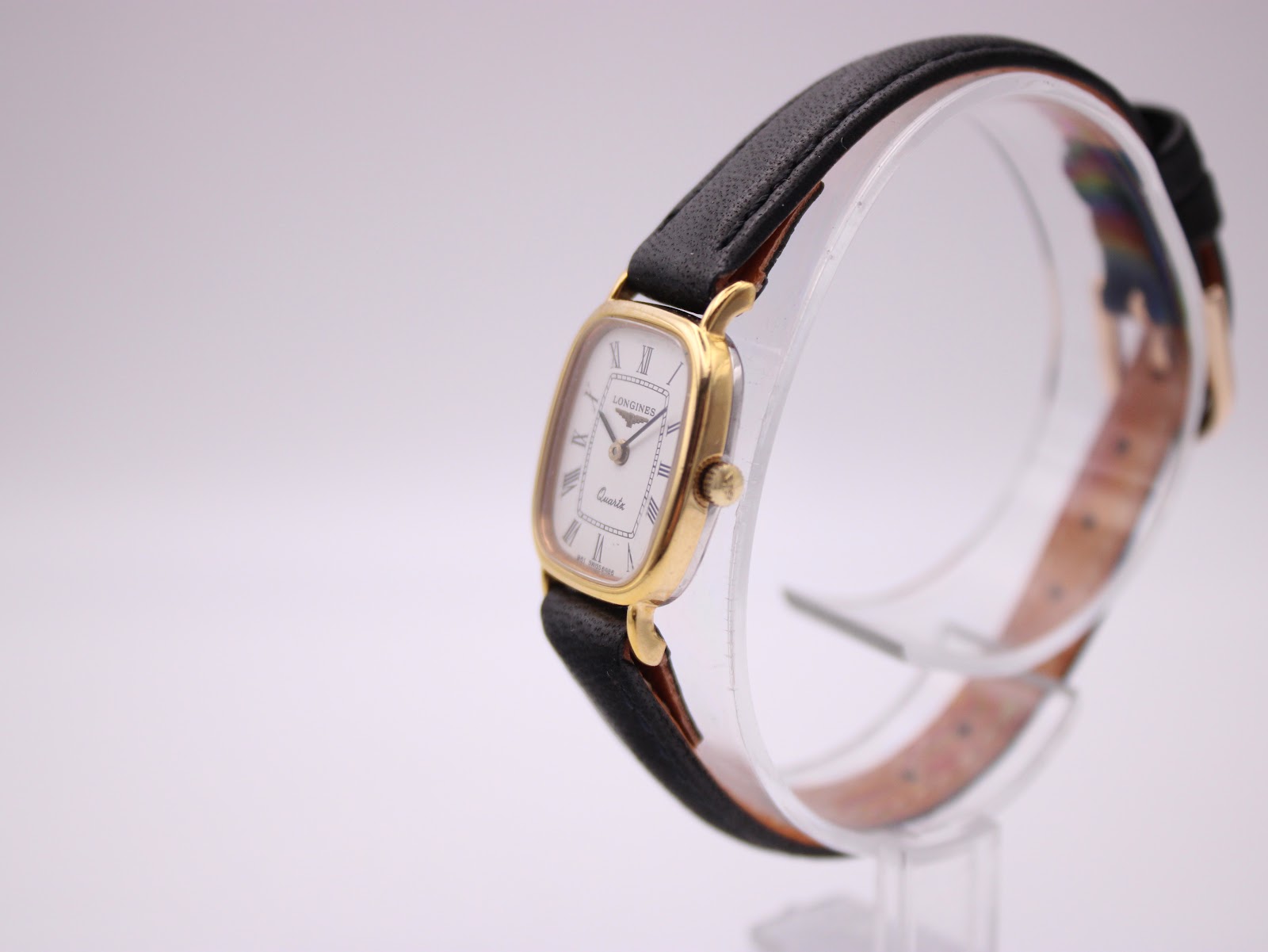 Angled side view of the Longines watch on a clear stand, showing the gold case and black leather strap.