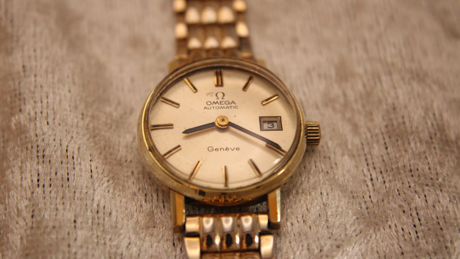 Direct front view of the Omega Geneve automatic watch with champagne dial and gold-tone bracelet on a textured background.
