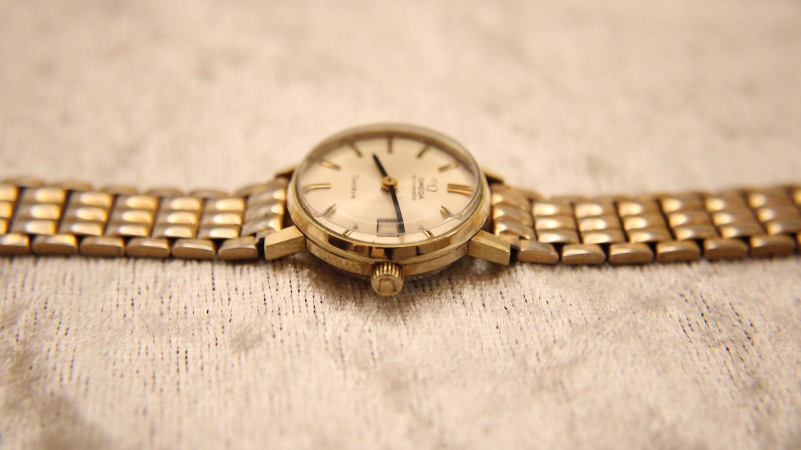 Side profile of the Omega Geneve watch showing the gold-plated case and bracelet on a light textured surface.