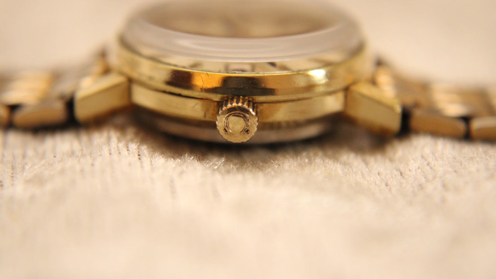 Close-up side view of the watch crown and gold-plated case on a textured background.