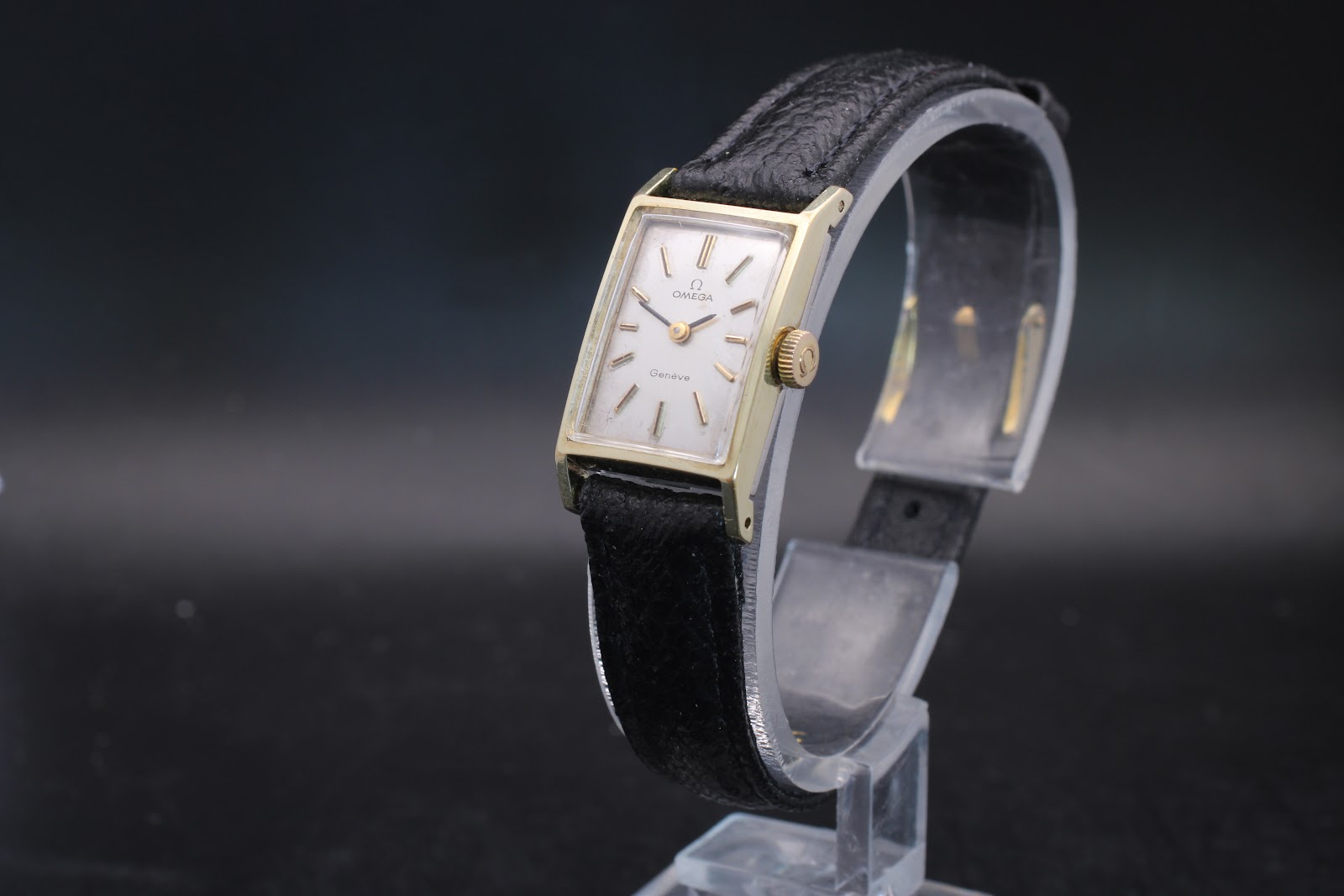 Side view of the vintage Omega Genève watch on a display stand, showing the gold rectangular case and black leather strap.