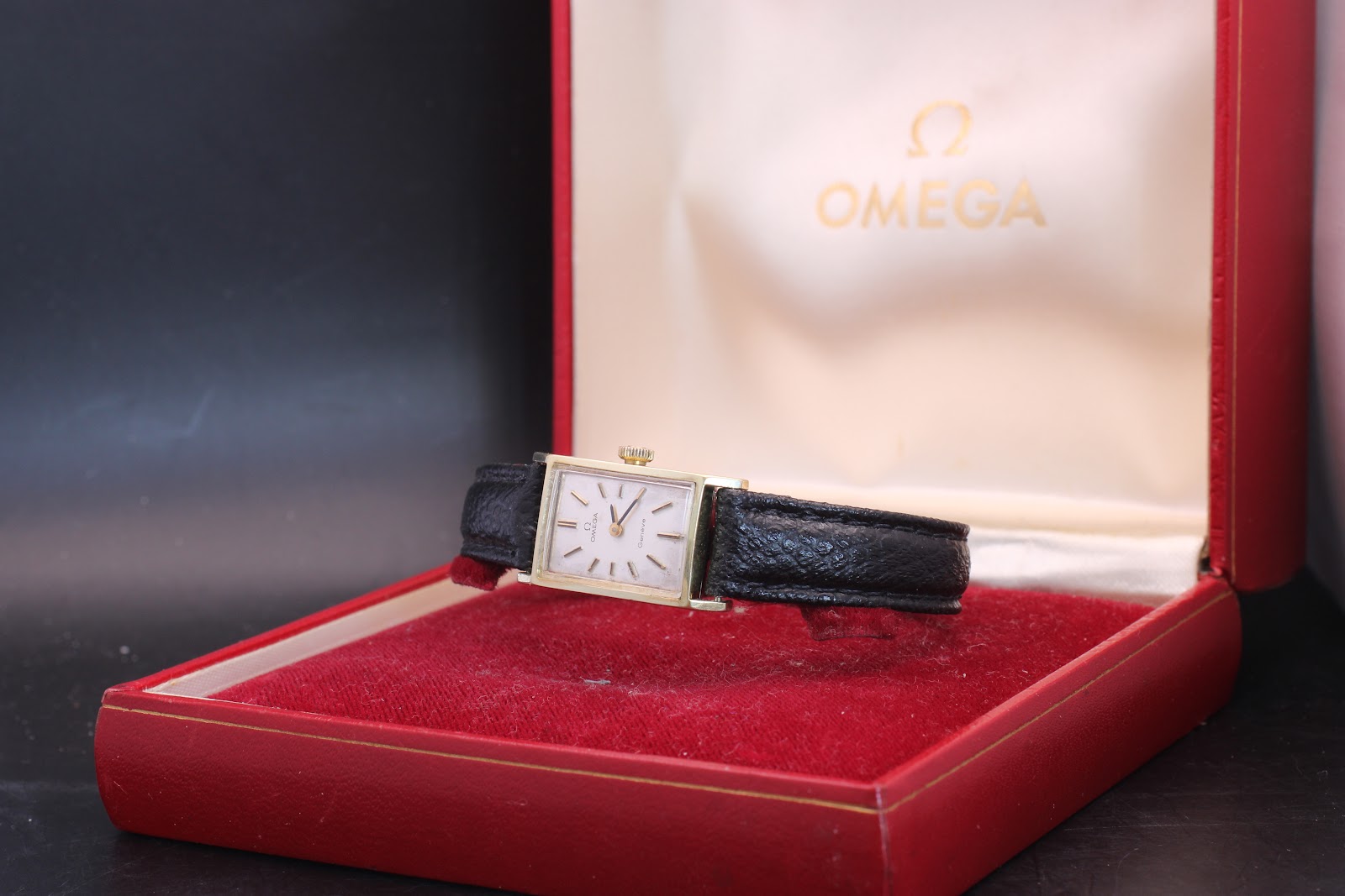 The vintage Omega Genève watch displayed open in its red original Omega presentation box, showing the watch face and strap.