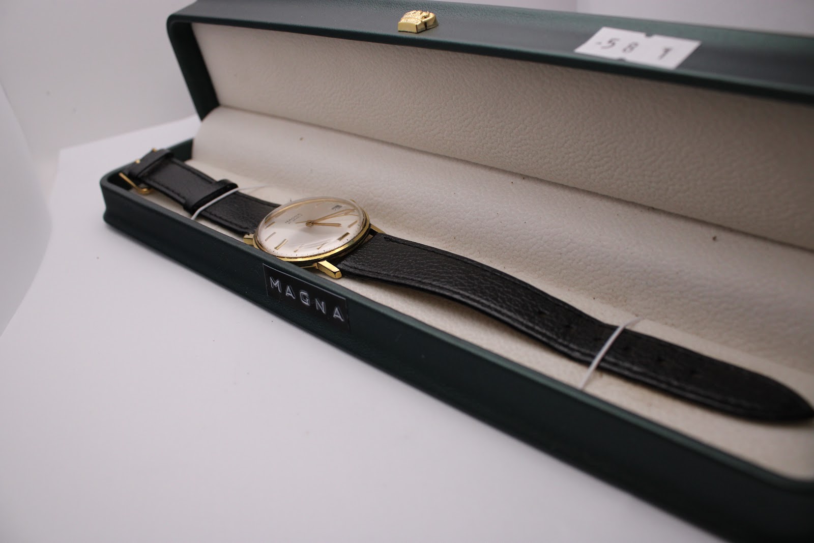 The Magna 21 jewels watch with its black leather strap, presented inside a dark green watch box, viewed from an elevated angle.