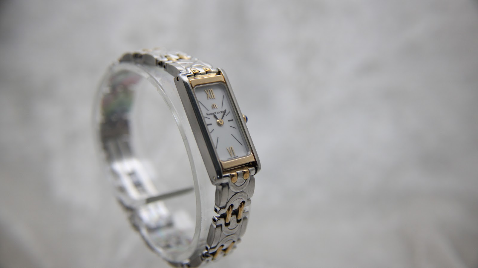 Three-quarter front view of the Maurice Lacroix watch on a clear stand, displaying the white dial with Roman numerals and the intricate two-tone bracelet.