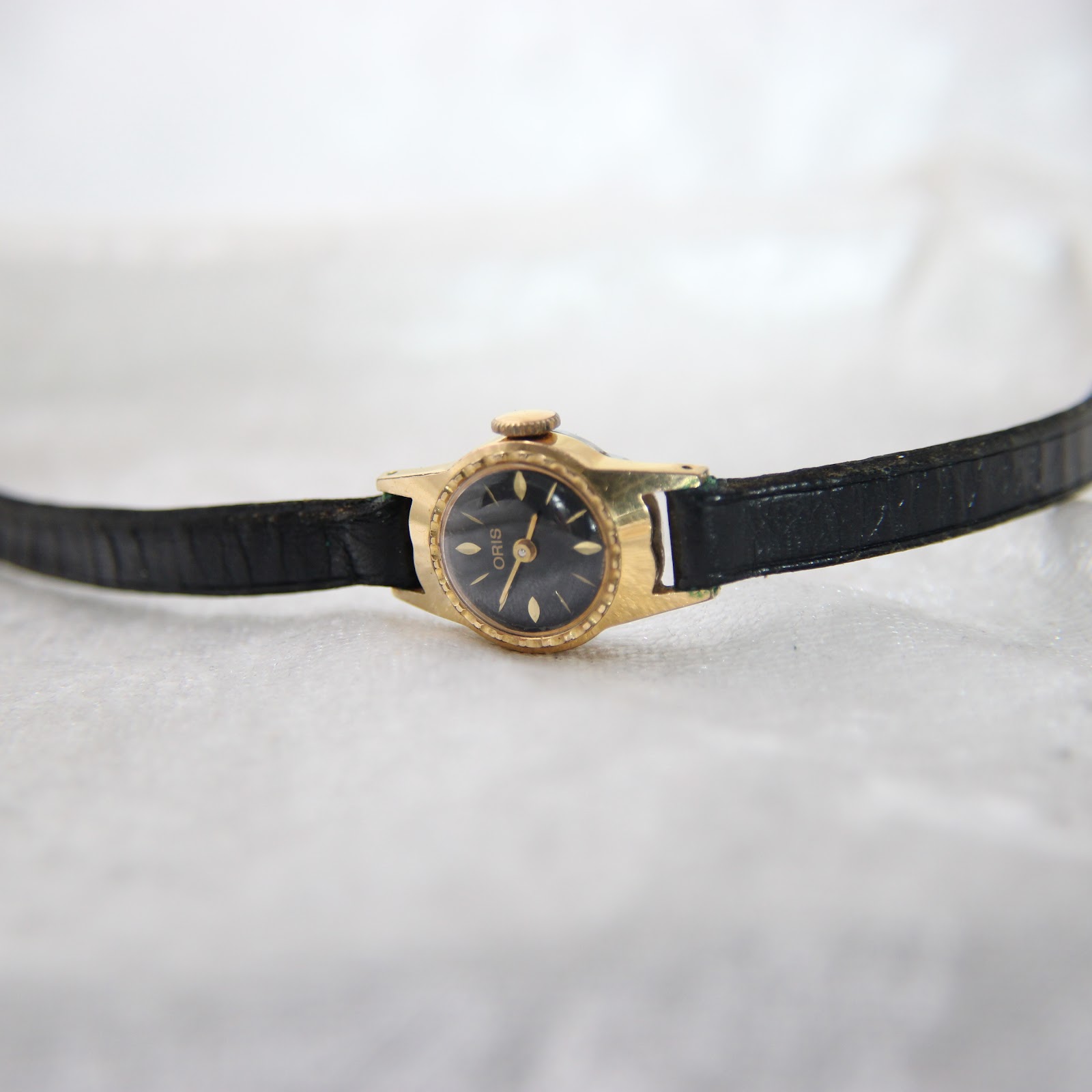 Top-down view of the gold plated Oris watch with its black leather strap laid flat on a white textured surface.