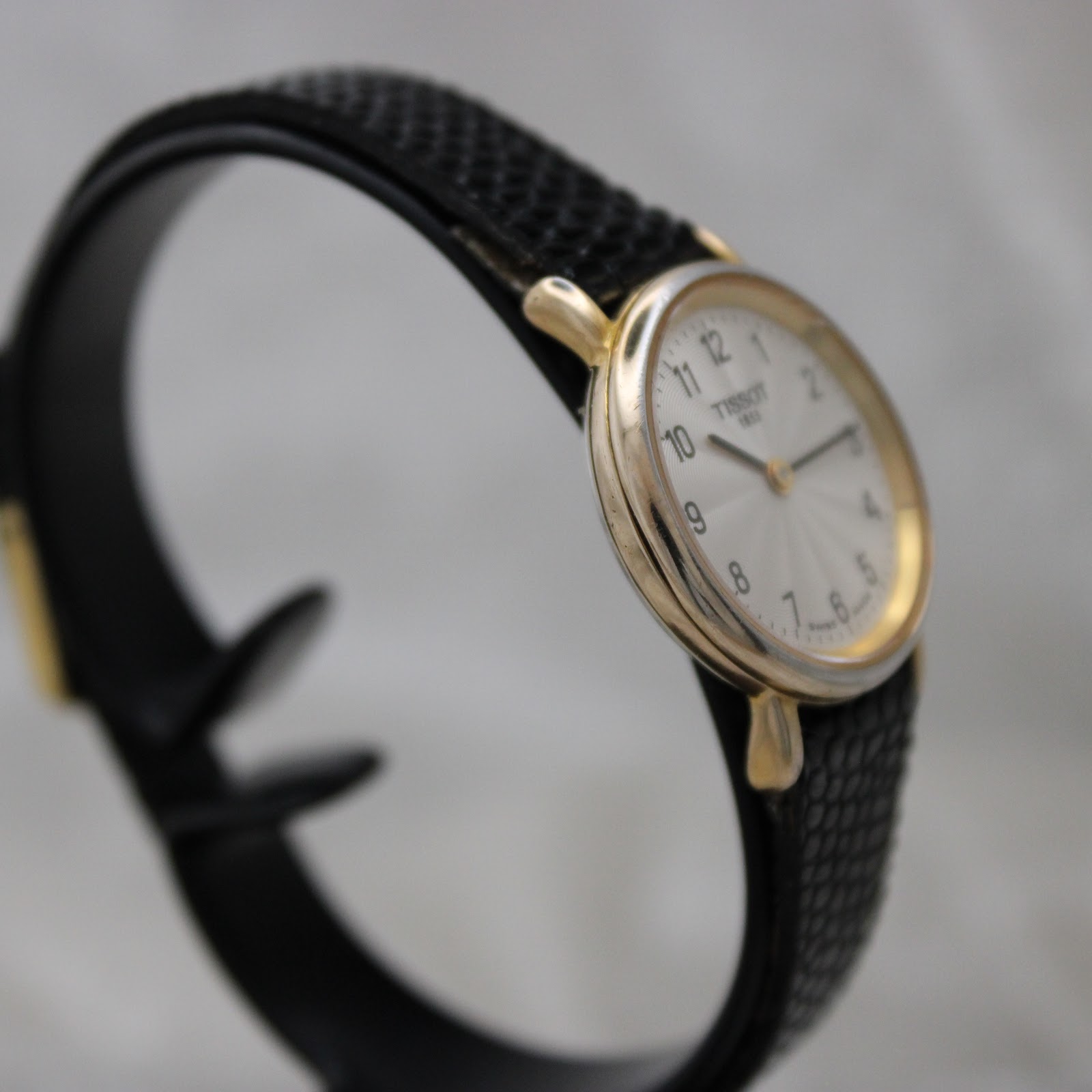 Angled view of the Tissot T852K watch from the left, showing the side profile of the gold-tone case and black leather strap on a watch stand.