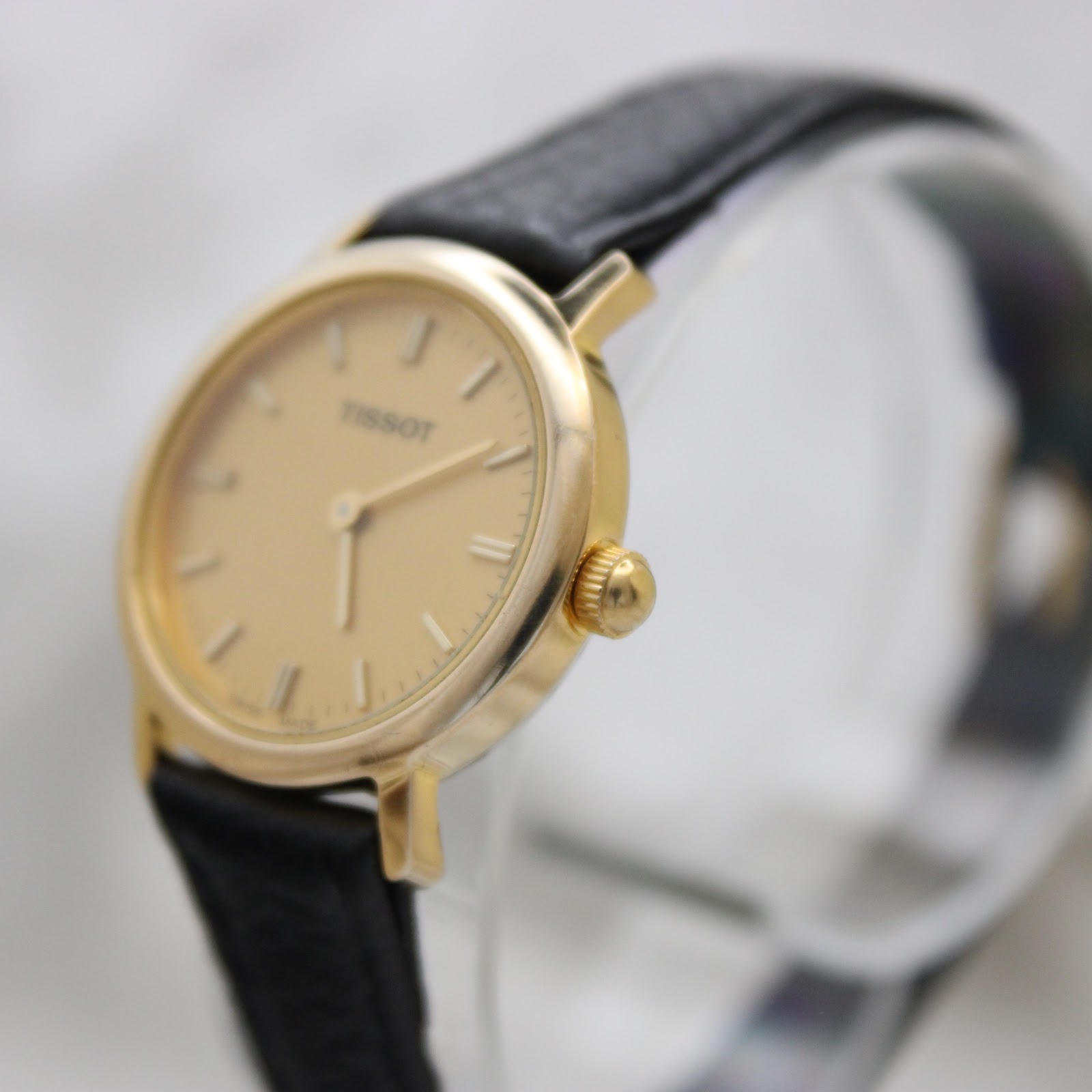 Angled view of the Tissot watch, showing the gold-plated case, crown, champagne dial, and black leather strap from the right side.