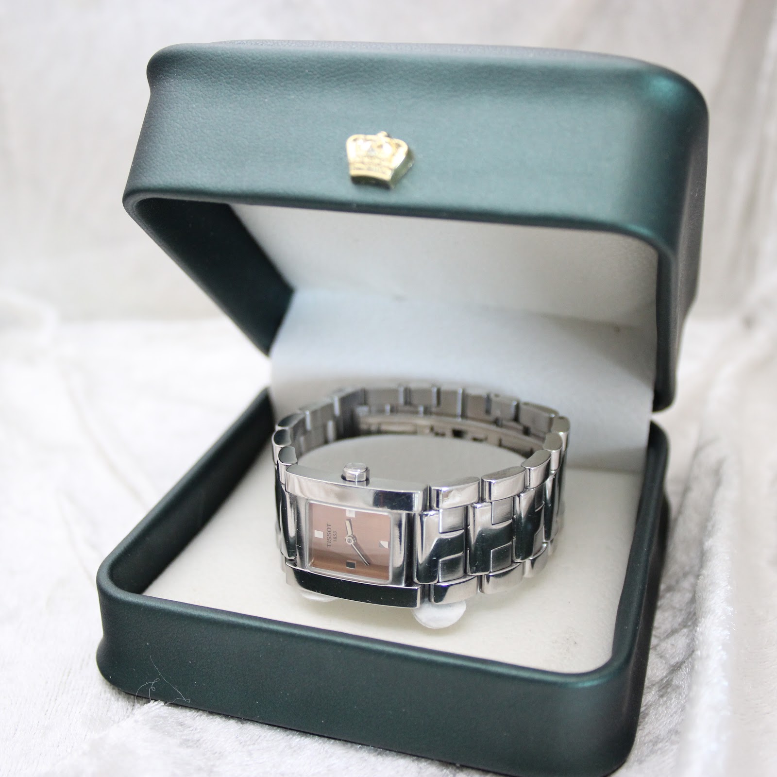 The Tissot square watch resting in an open green presentation box with a golden crown emblem, though the box is not included with the sale.