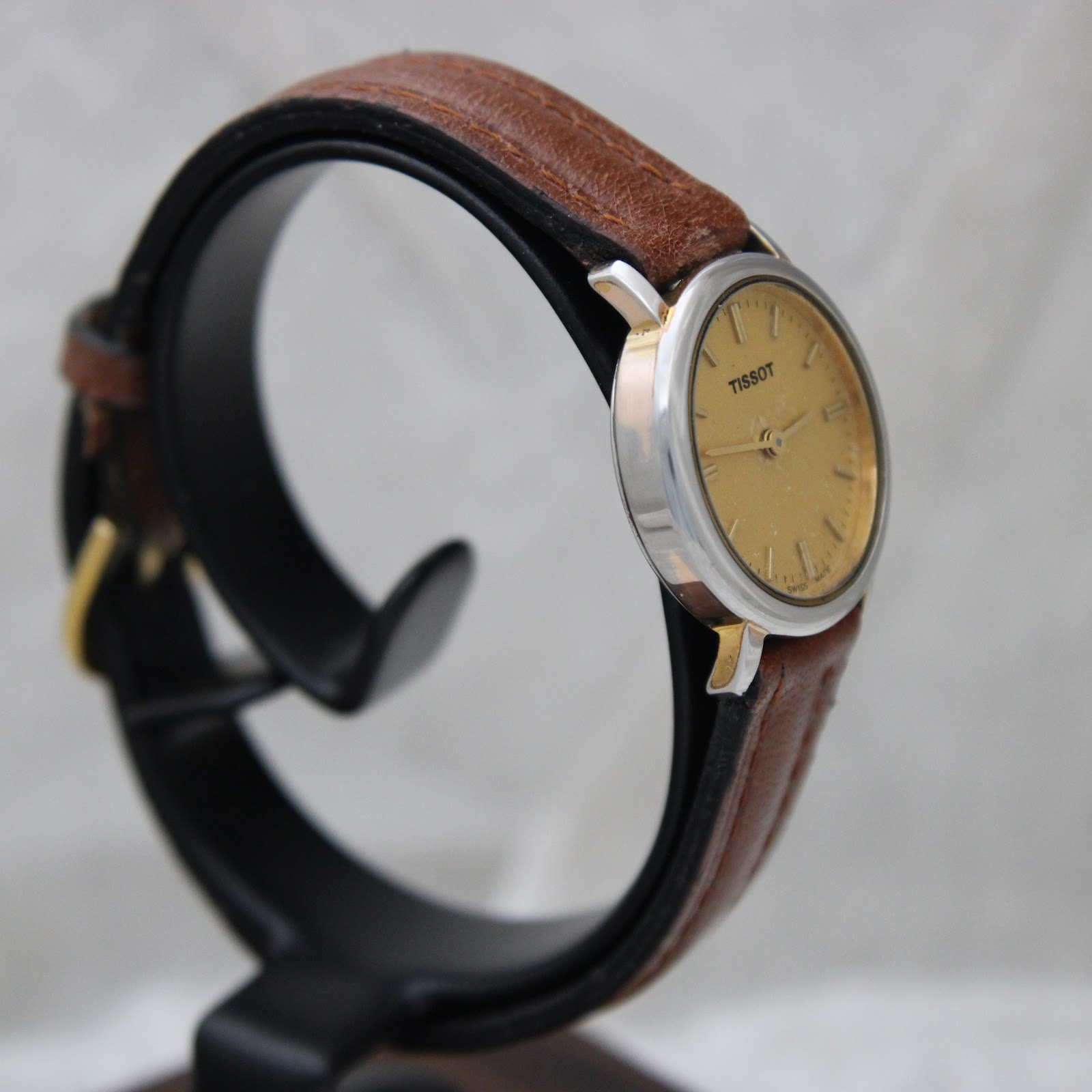 Right side view of the Tissot watch on a stand, showing the silver-tone case and gold-tone lugs, with the brown leather strap wrapped around. The dial is angled away.