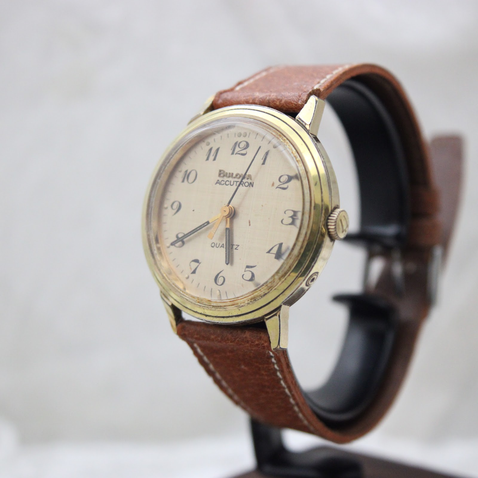 Angled side view of the Bulova Accutron watch on a display stand, showing the gold-plated case and brown leather strap.