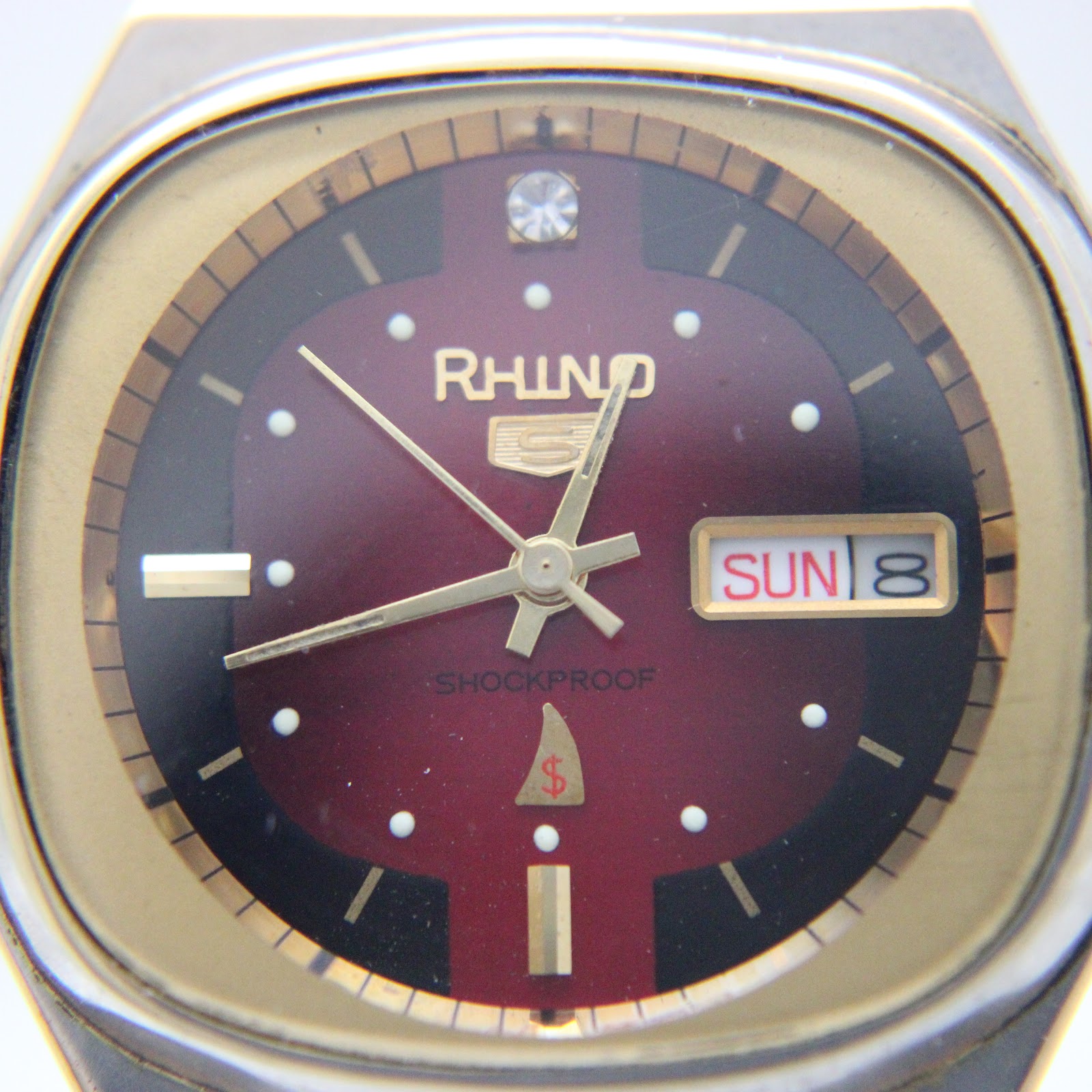 Macro close-up of the Rhino watch dial, revealing the luminous hands, applied indices, diamond-style 12 o’clock marker, day/date window, 'shockproof' text, and sharkfin logo.